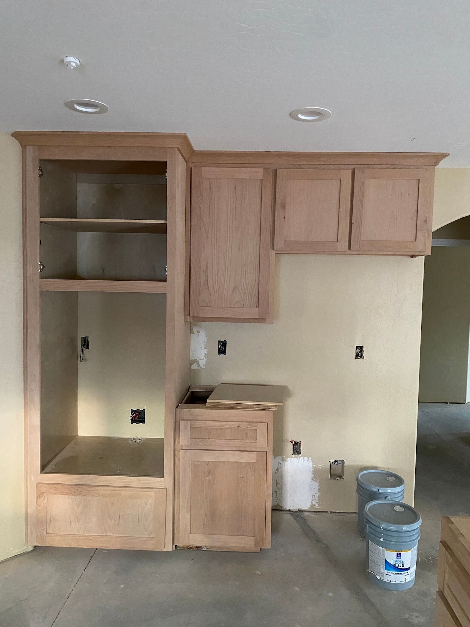 Kitchen cabinetry under construction with open and closed wooden cabinets, electrical outlets, and two buckets on the floor.