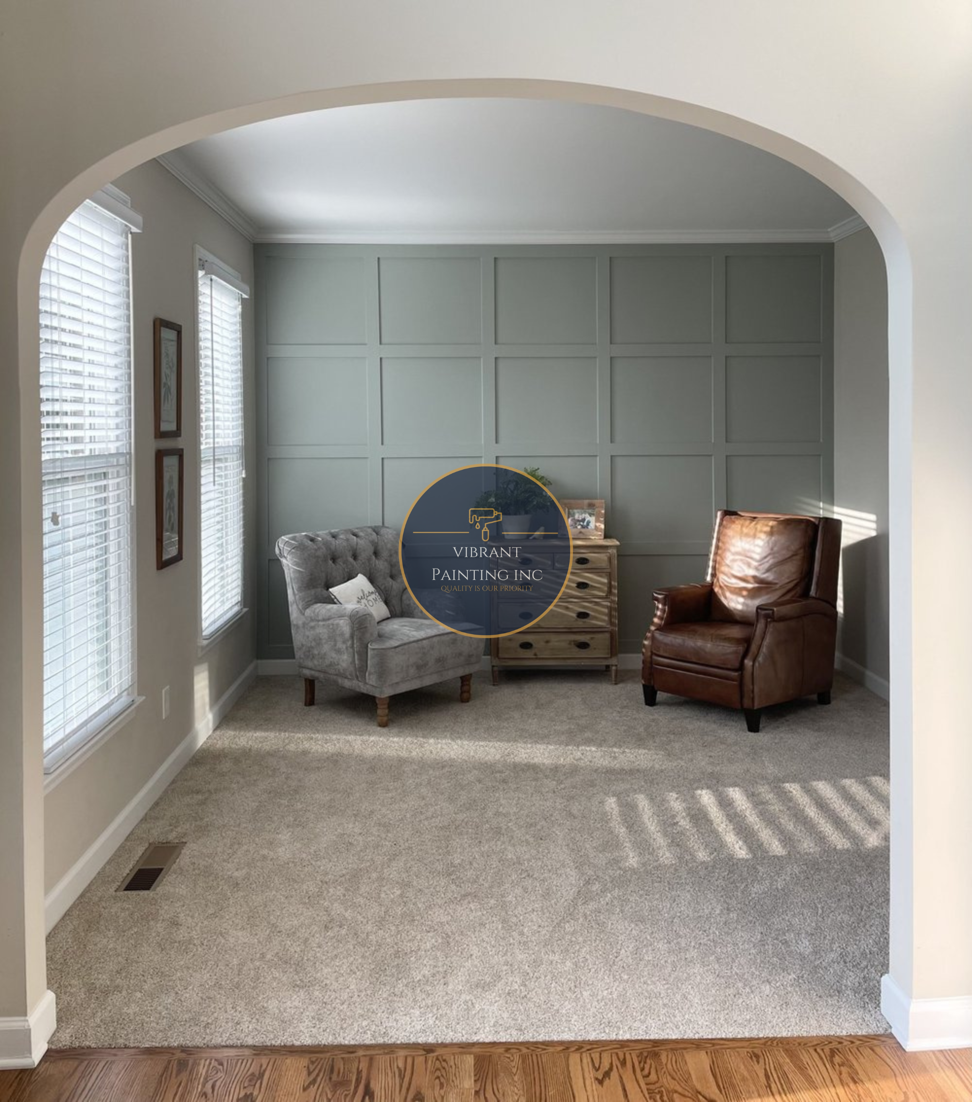 Living room with light beige carpet, gray accent wall, two windows with blinds, a tufted gray armchair with a pillow, a wooden side table, and a brown leather armchair.