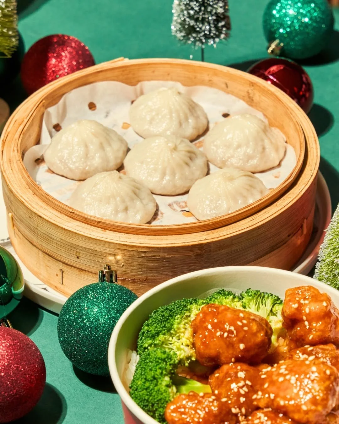 Steamed dumplings in a bamboo steamer basket and glazed chicken with broccoli in a white bowl, surrounded by Christmas ornaments.