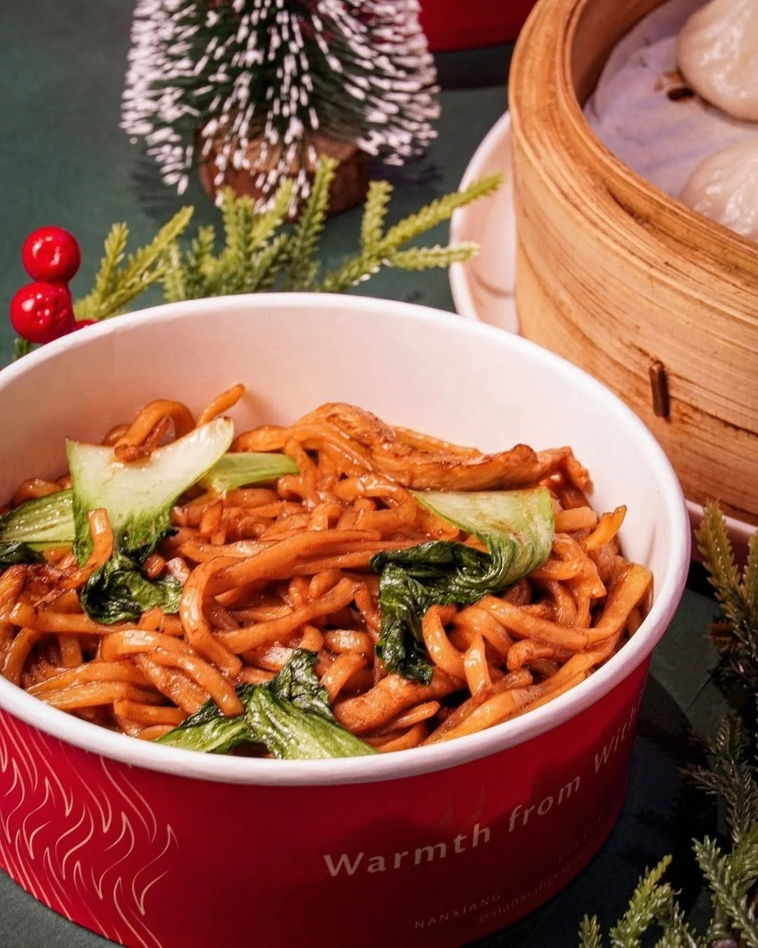 A bowl of stir-fried noodles with vegetables, including bok choy, on a festive table with Christmas decorations such as pine branches and red berries.