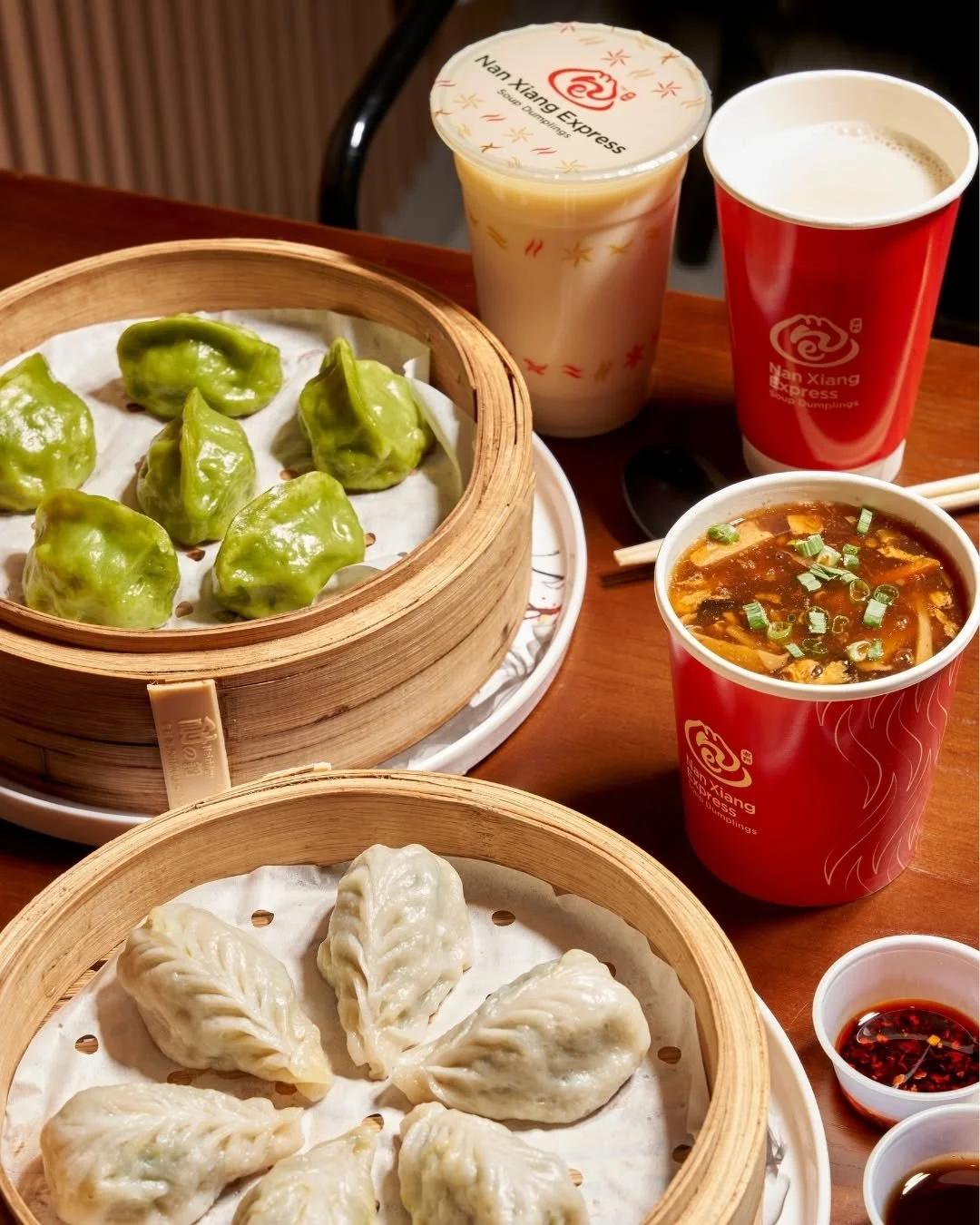 Steamed green and white dumplings on bamboo steamers with cups of soup, tea, and a beverage on a wooden table.