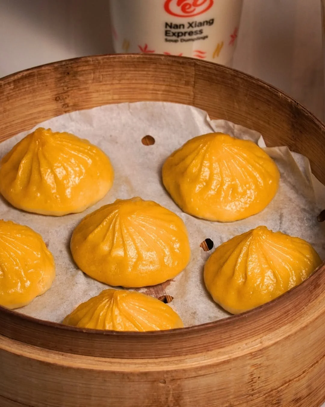 Six dim sum dumplings with yellow wrappers placed in a bamboo steamer basket over parchment paper, with a can of Coke Nan Xiang Express in the background.