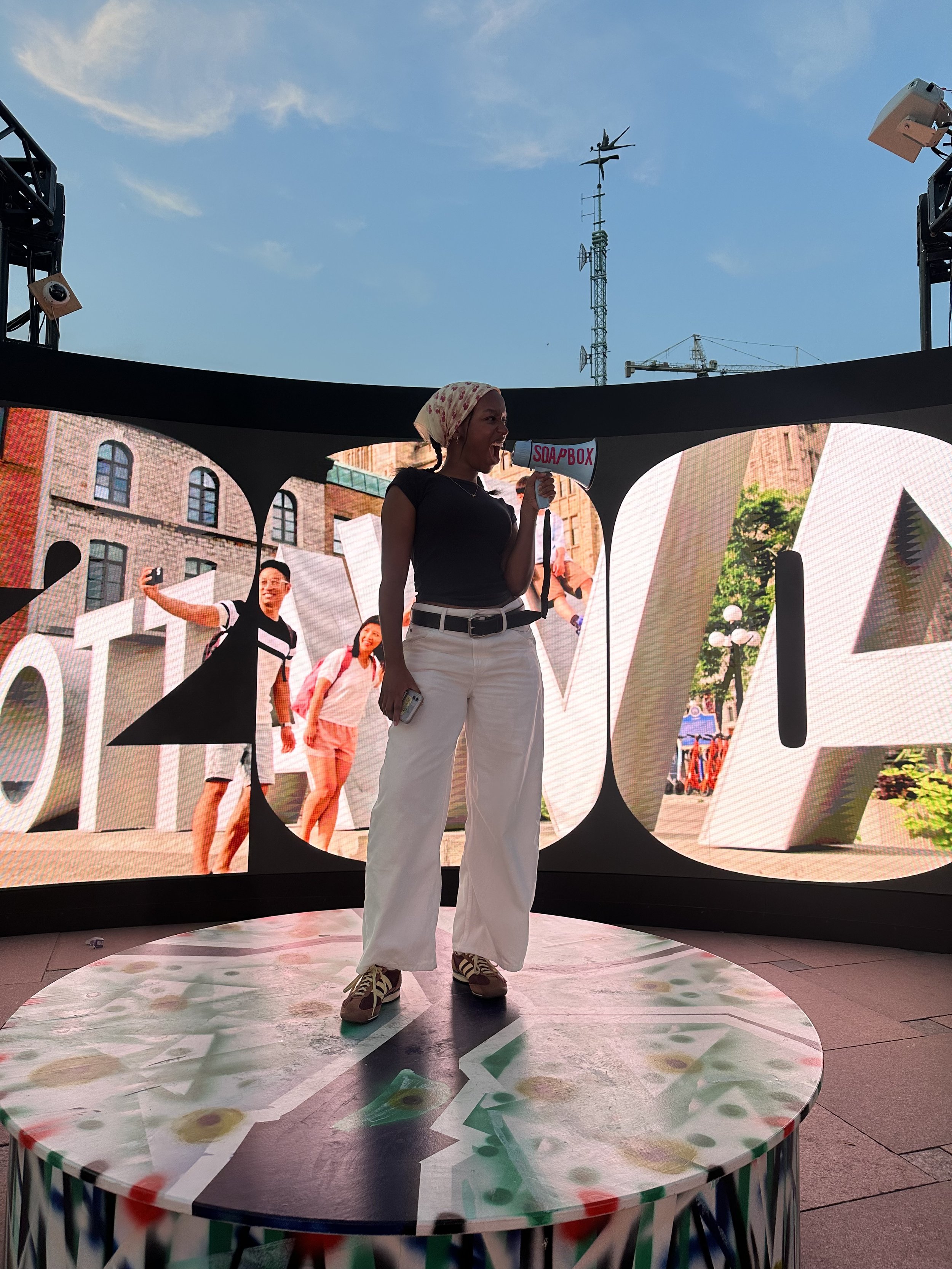 Person wearing a headscarf and white pants holding a megaphone on a stage with a large LED screen behind showing people in a city scene.