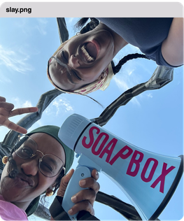 Two people taking a selfie outdoors with a soap box in the background labeled 'SOAP BOX'. The sky is clear and blue.