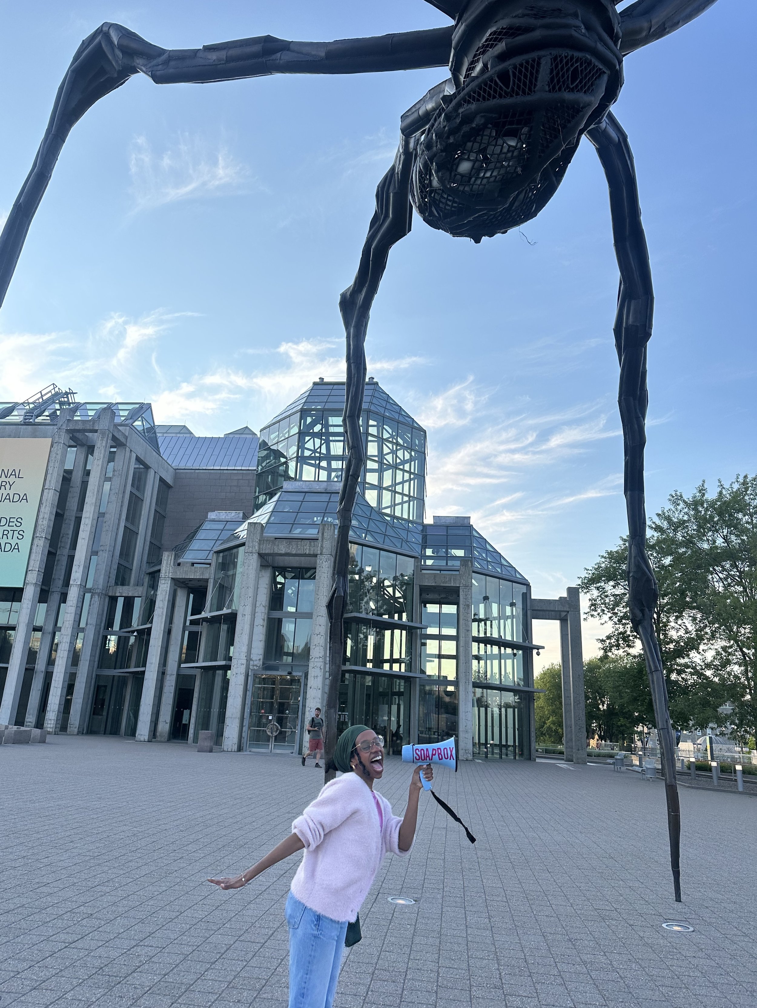 A woman holding a sign that says 'SOAPBOX' and posing playfully in front of a modern glass building, with a large black spider sculpture overhead and other people walking in the background.