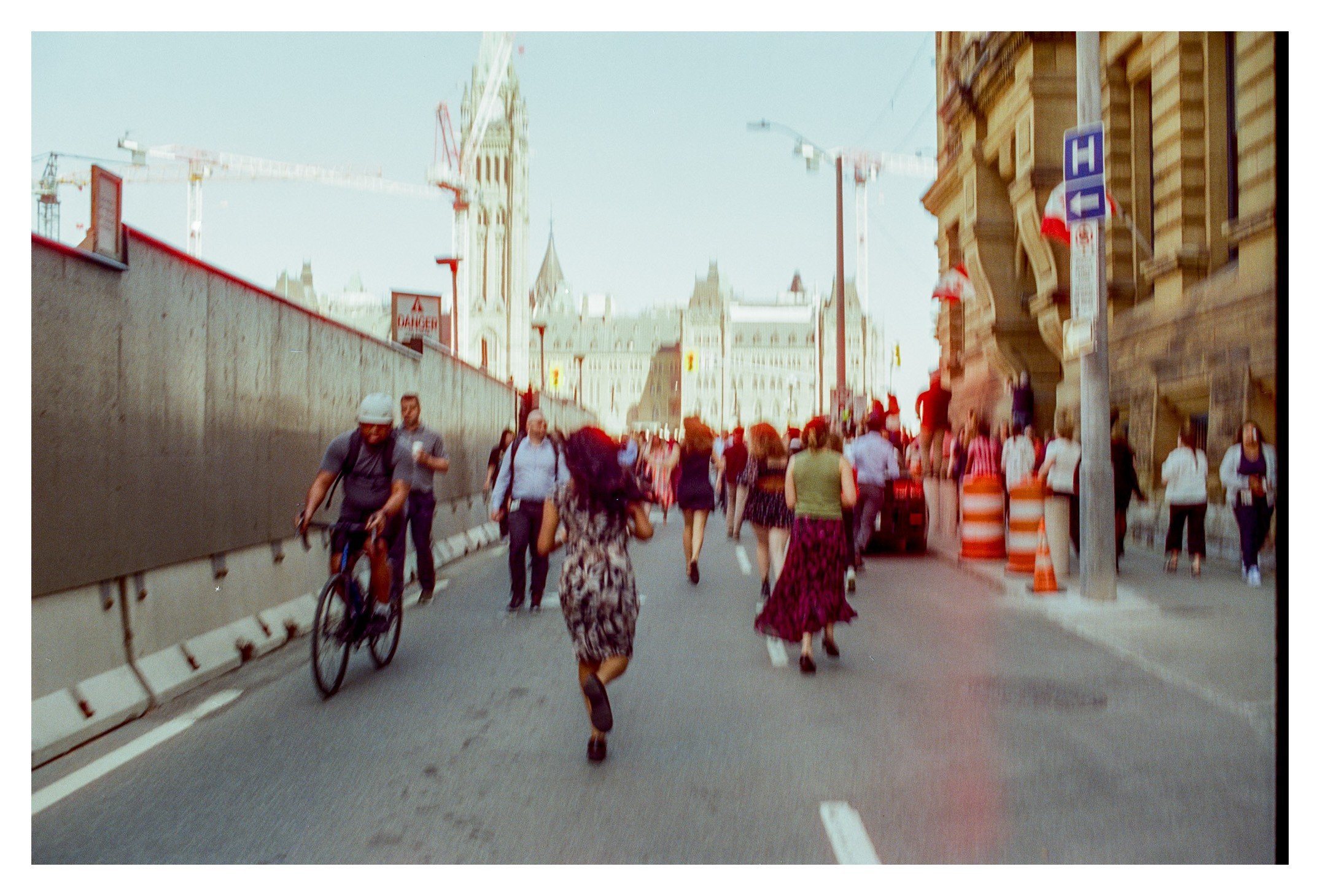 People walking and cycling on a city street near Parliament Hill, with construction cranes and historic buildings visible in the background.
