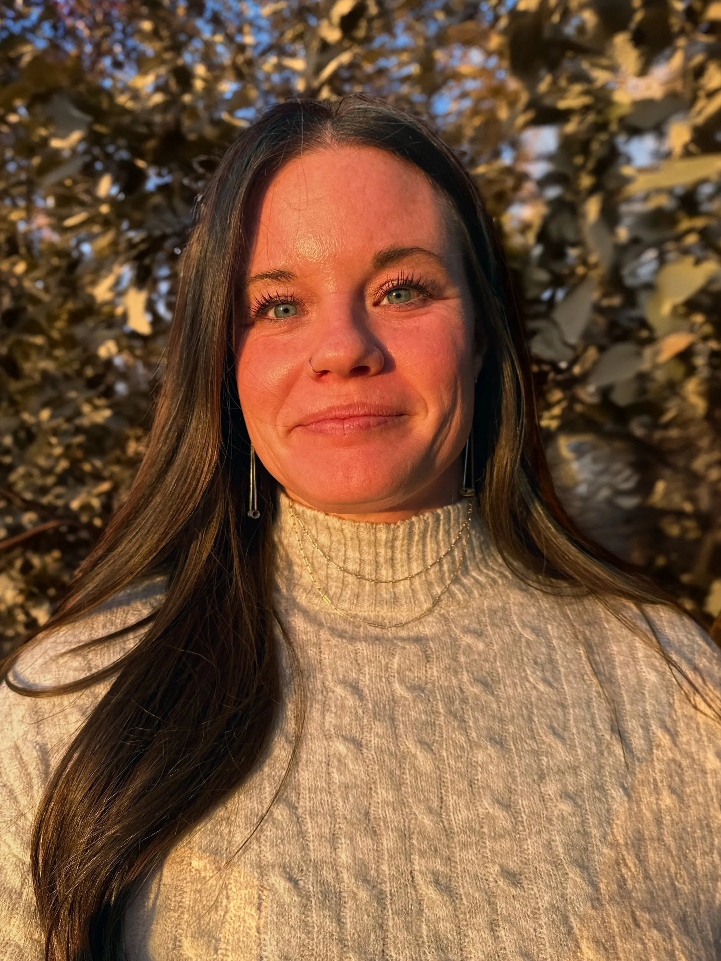 A woman with long dark hair and light-colored eyes, wearing a beige turtleneck sweater, standing outdoors in sunlight with a background of dense bushes or trees.