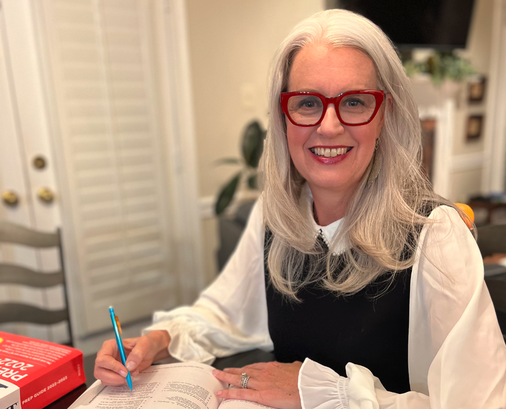 The ACT Mom with long gray hair, wearing red glasses, a white blouse, and a black vest, smiling while sitting at a table with an open textbook, a blue pen in her hand, and a red ACT prep book nearby in a cozy room.