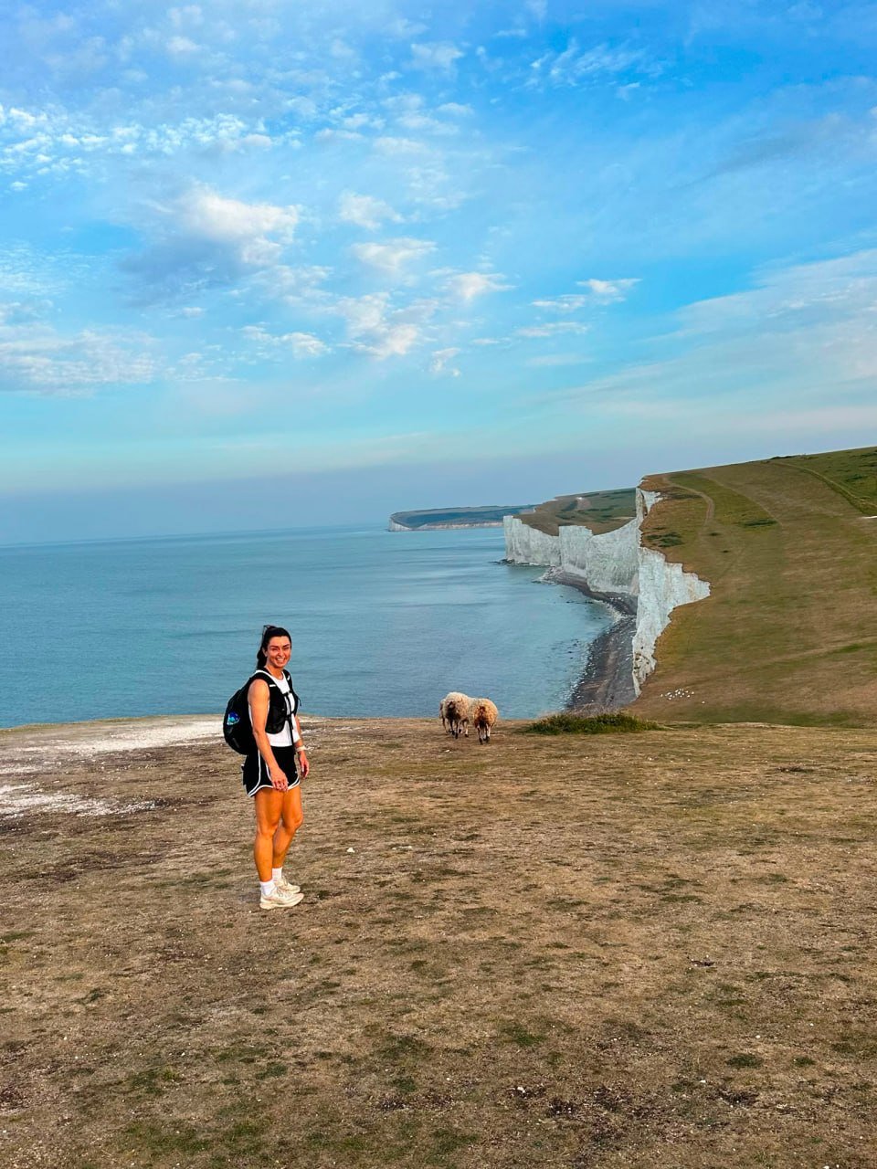 A woman with a backpack standing on a grassy cliff overlooking the ocean with white chalk cliffs in the background, accompanied by sheep in the distance.