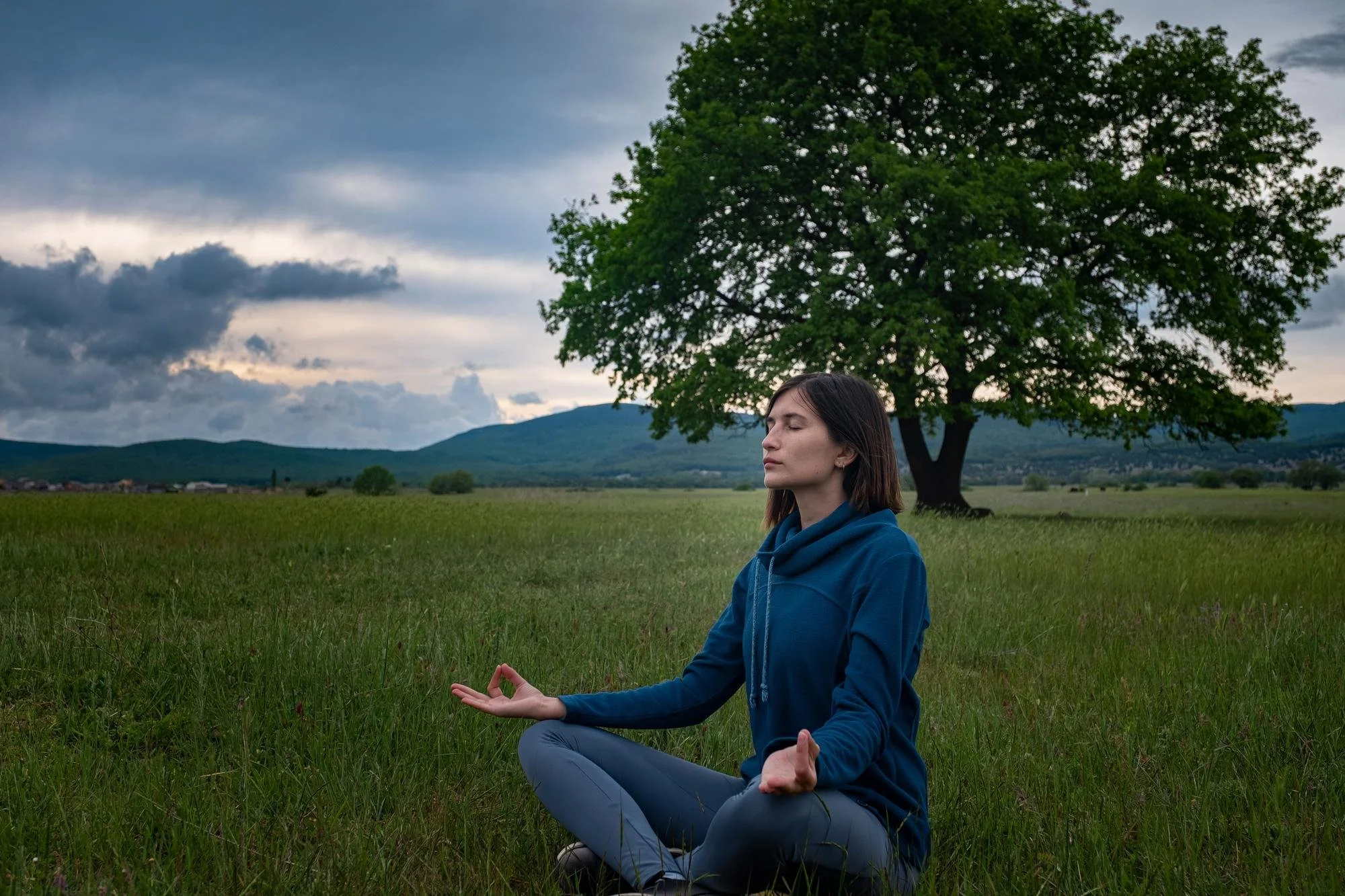 A woman with dark brown hair sitting cross-legged in a meditation pose outdoors, with her eyes closed, under a large green tree with a scenic background of mountains and cloudy sky.