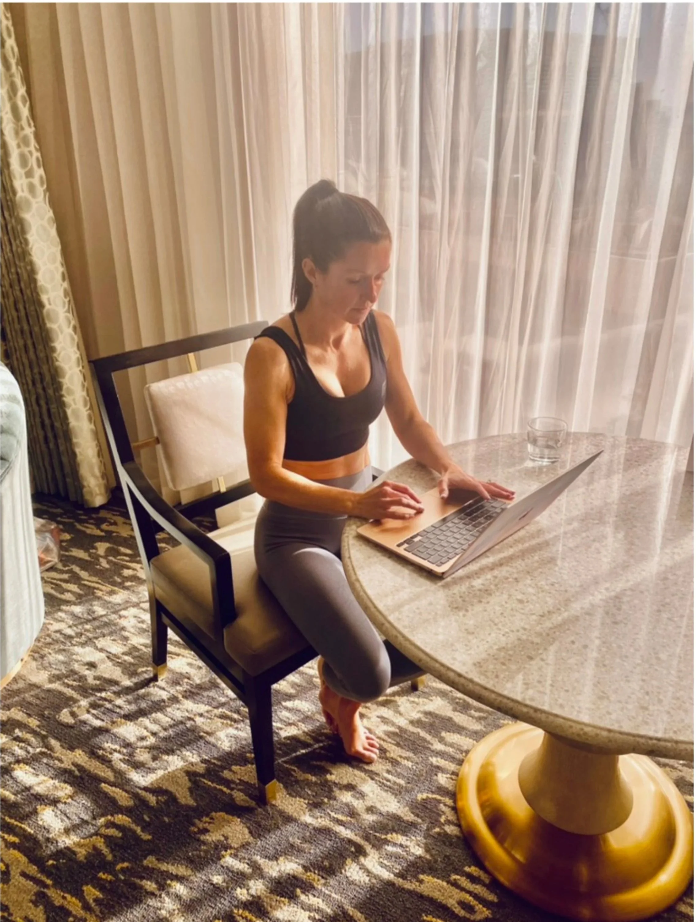 A woman in workout attire sitting at a round table using a laptop with a glass of water nearby, in a room with patterned carpet and sheer curtains.