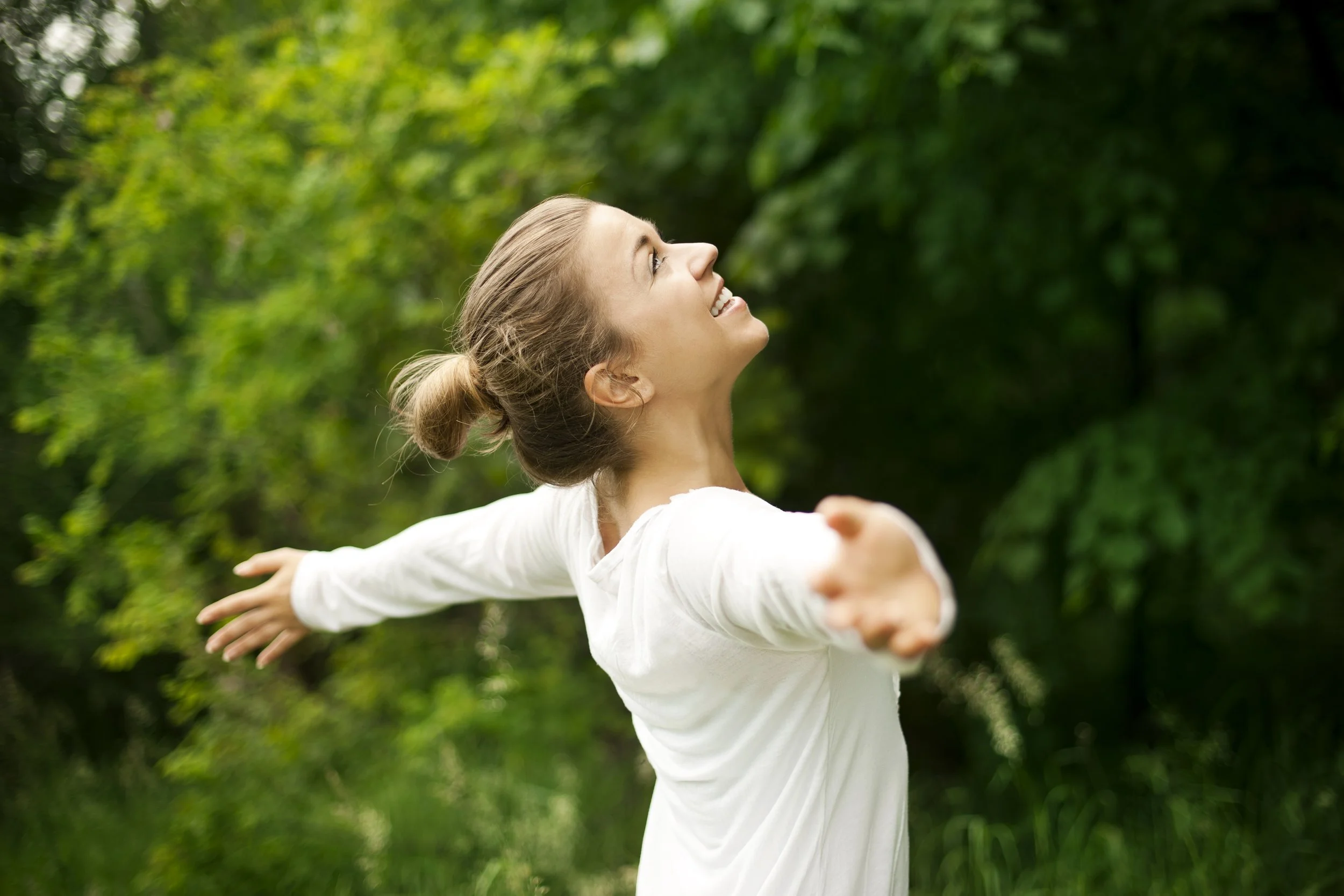 A woman with blonde hair tied back, wearing a white shirt, smiling with arms outstretched, outdoors in a green park.