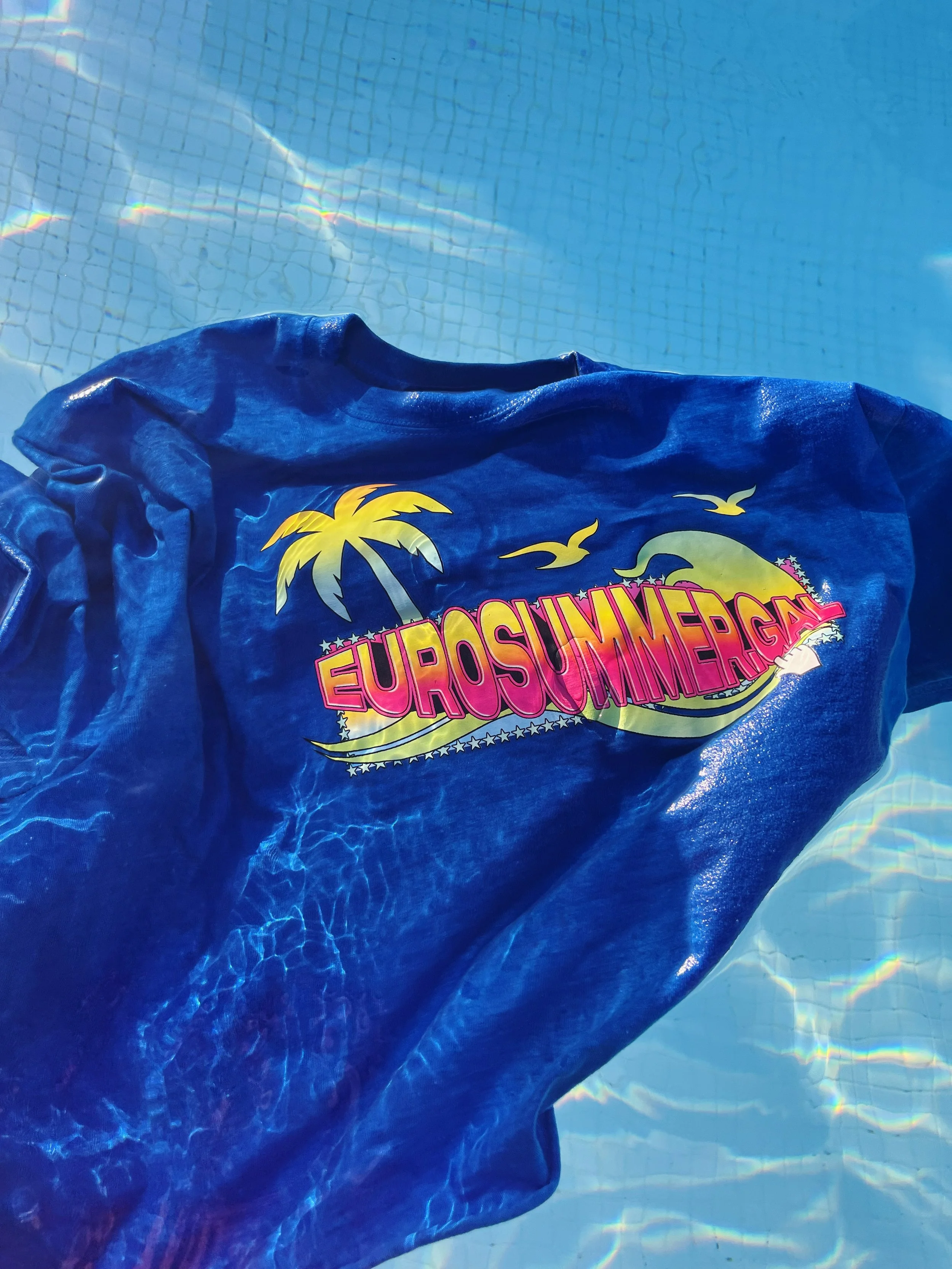 Camiseta azul con logotipo colorido de EuroSummerPG con un sol, palmeras y gaviotas, parcialmente mojada, en una piscina con reflejos en el agua.