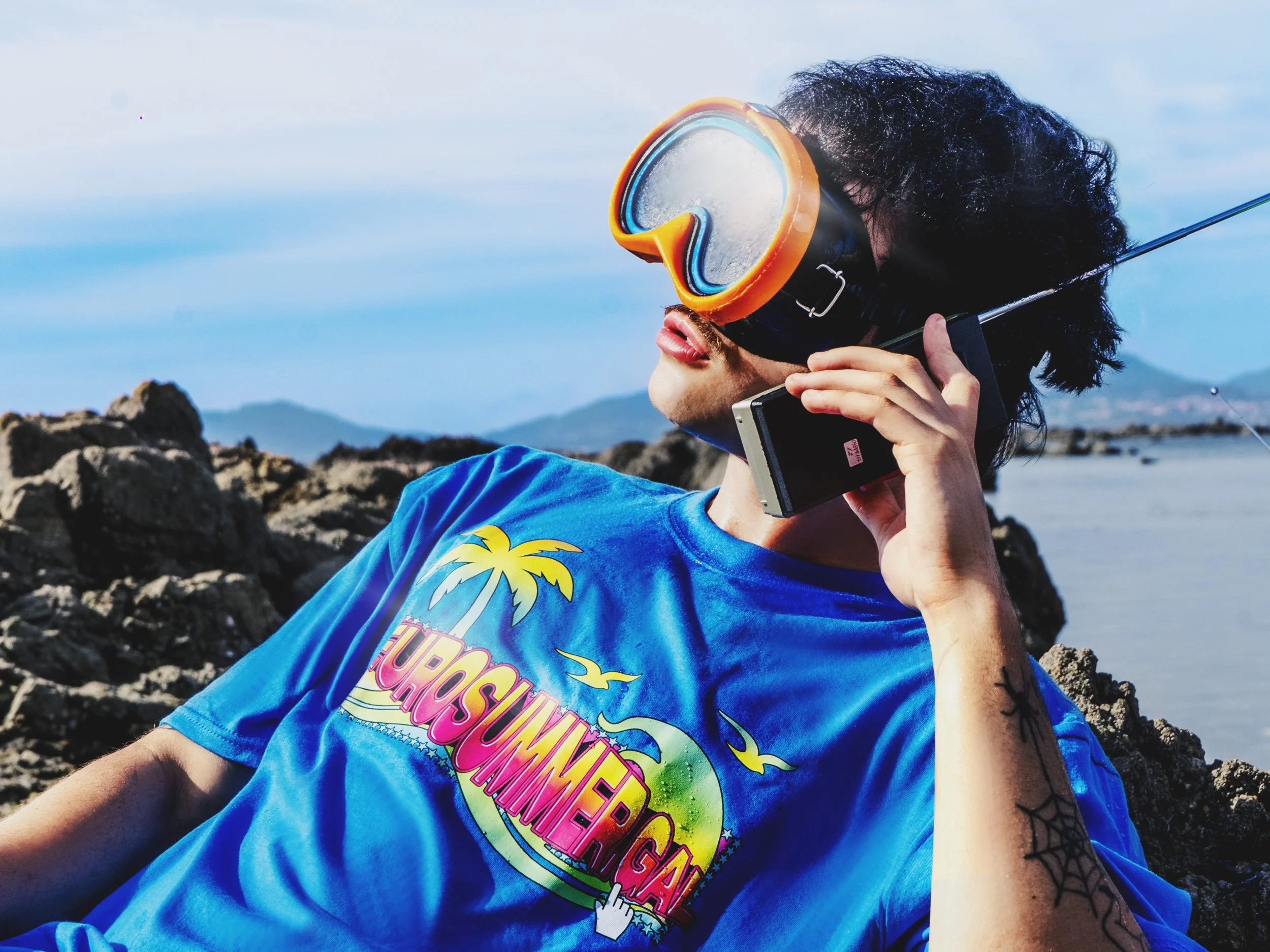 Persona con gafas de snorkel y traje azul, descansando en una roca junto al mar con montañas al fondo.