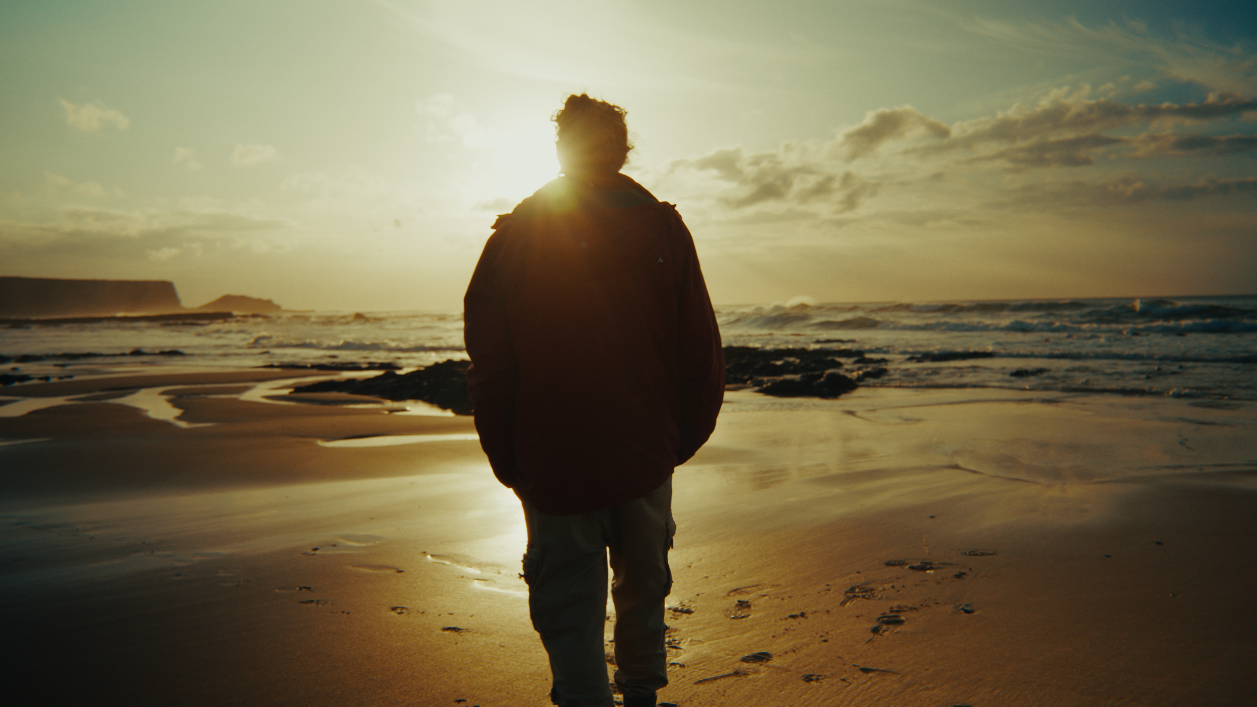 Person walking on a beach at sunset with the sun behind them, ocean waves, and cloudy sky in the background.