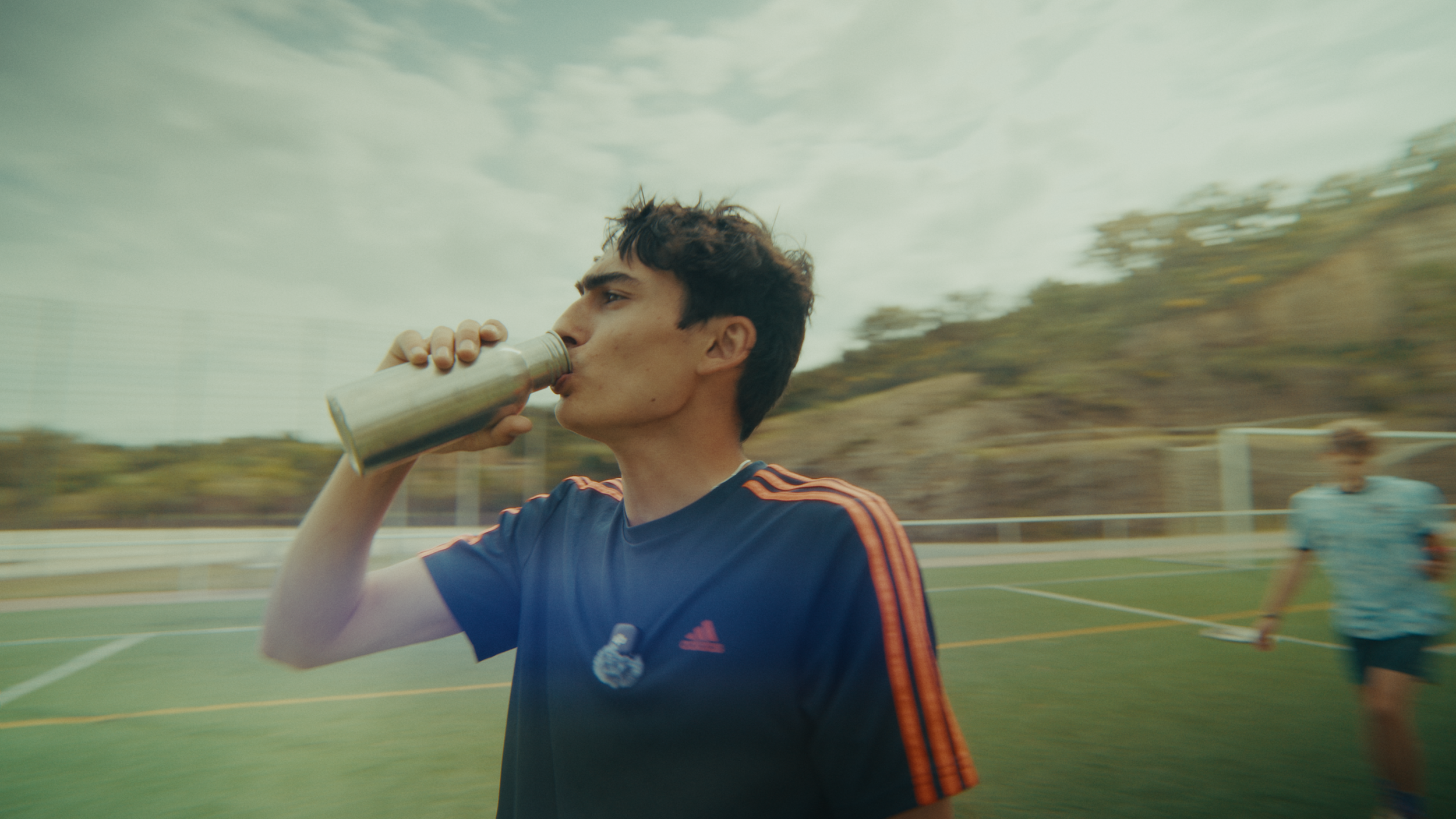 A young man in a blue sports jersey drinking from a silver water bottle on a sports field, with another person in the background and cloudy sky overhead.