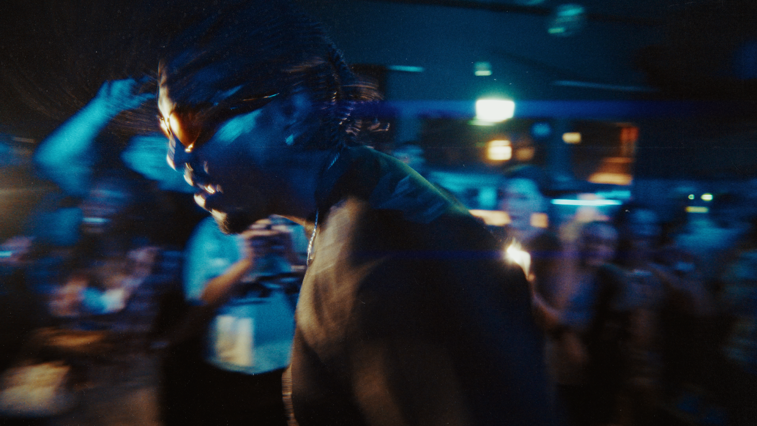 A person with sunglasses and shoulder-length hair dancing in a dark, crowded nightclub with colorful lights.