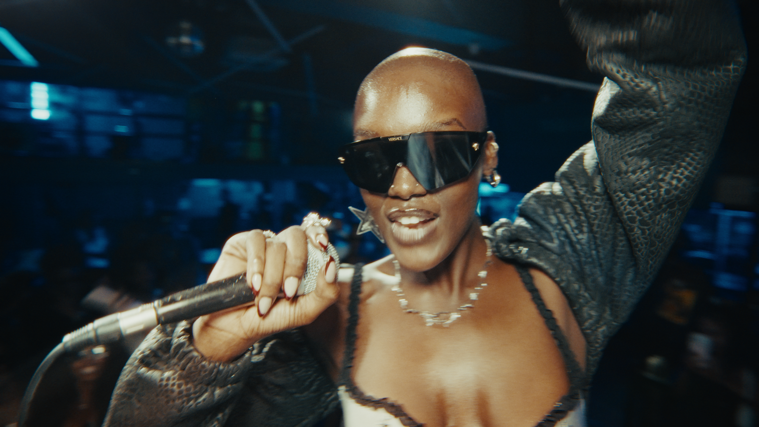 A woman with a buzz cut dark sunglasses, holding a microphone, smiling in a nightclub setting with blue lighting.