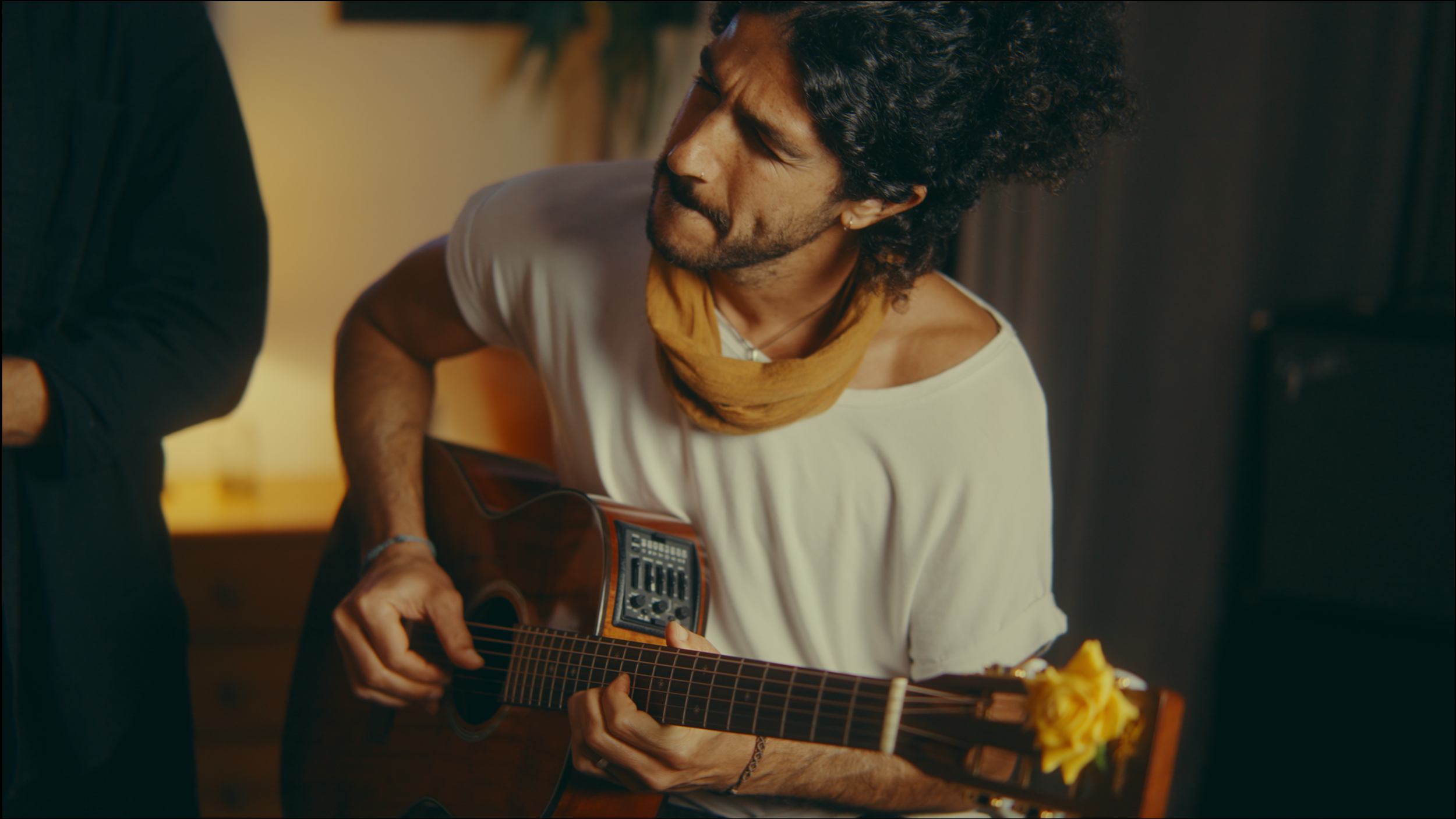 A man with curly hair wearing a white t-shirt and a yellow scarf playing an acoustic guitar indoors.