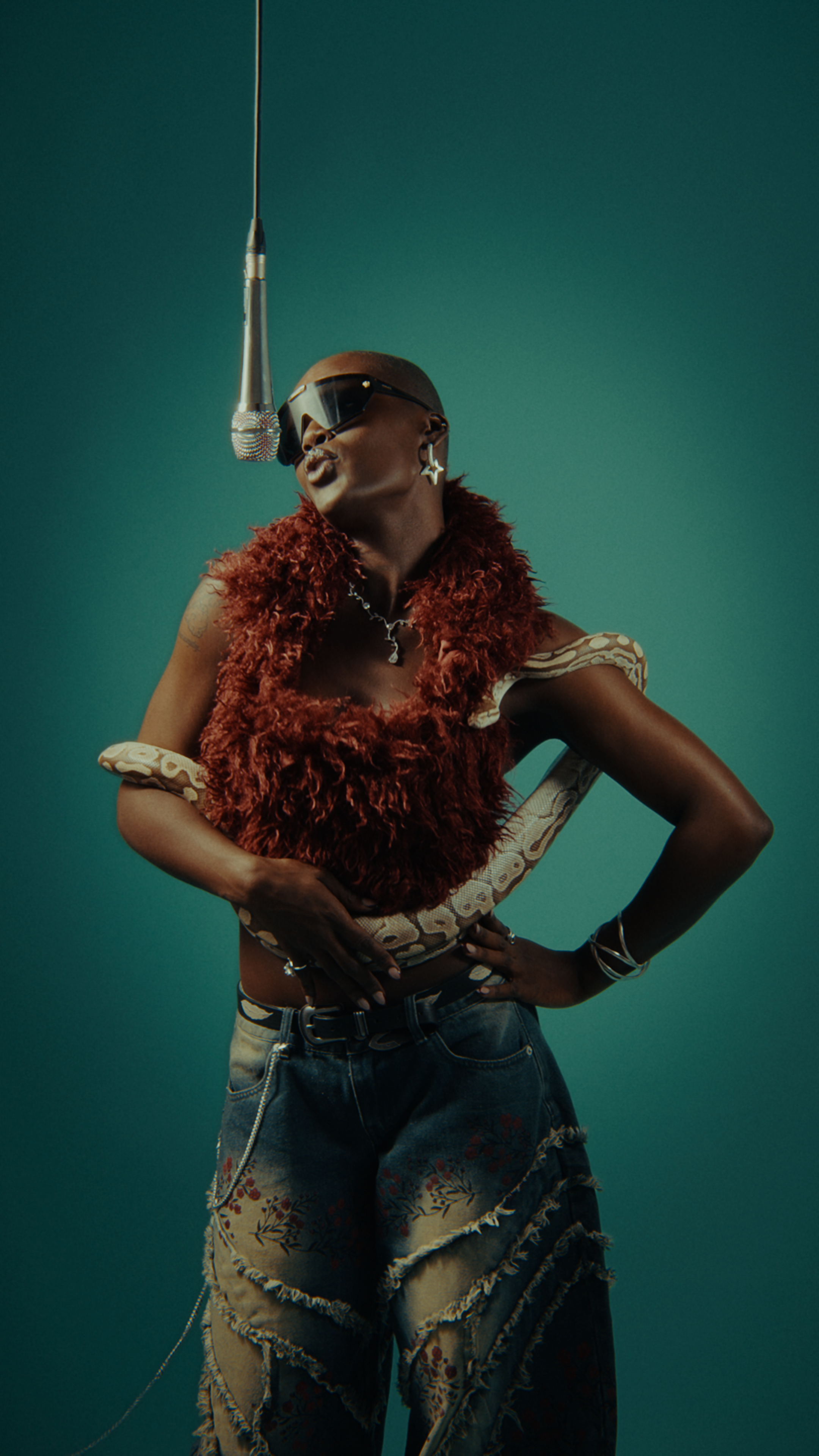 A person wearing dark glasses, a star earring, a necklace, a red furry vest, snake-print accessories, and baggy embroidered pants, standing against a teal background, in front of a hanging microphone.