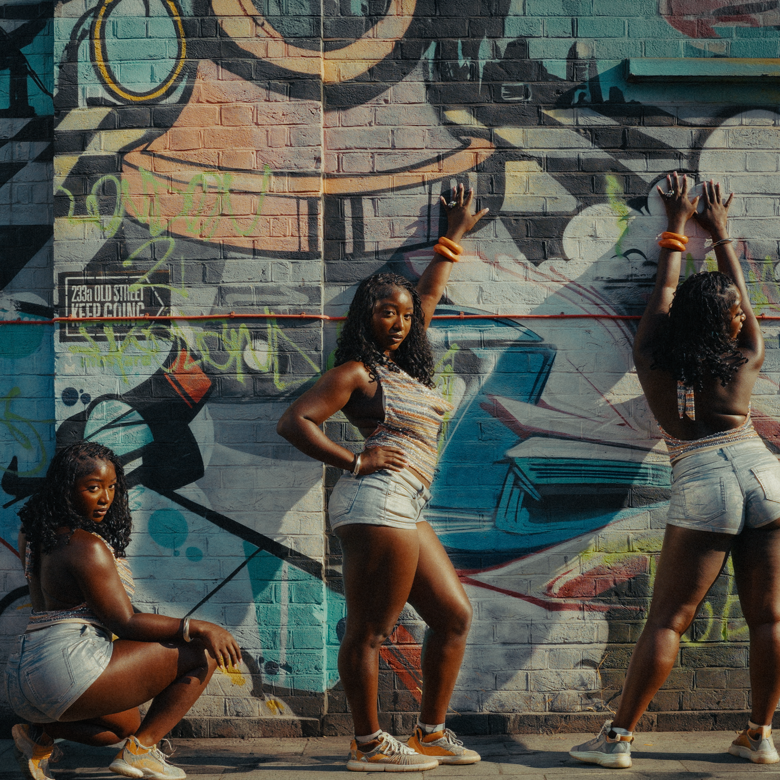 Group of women standing in front of a colorful graffiti wall, with three women visible, wearing casual summer clothing, and posing with hands on hips and arms raised.