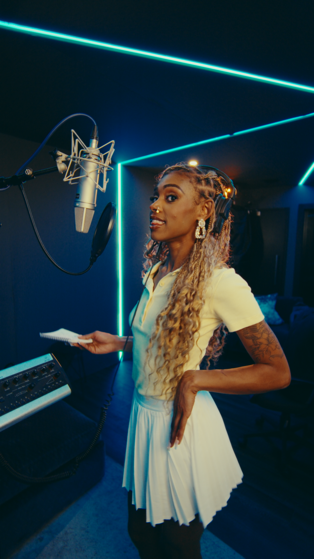 A woman with curly blonde hair and tattoos, wearing a yellow polo shirt and a white skirt, stands in a recording studio with a microphone and audio equipment, illuminated by blue neon lights.