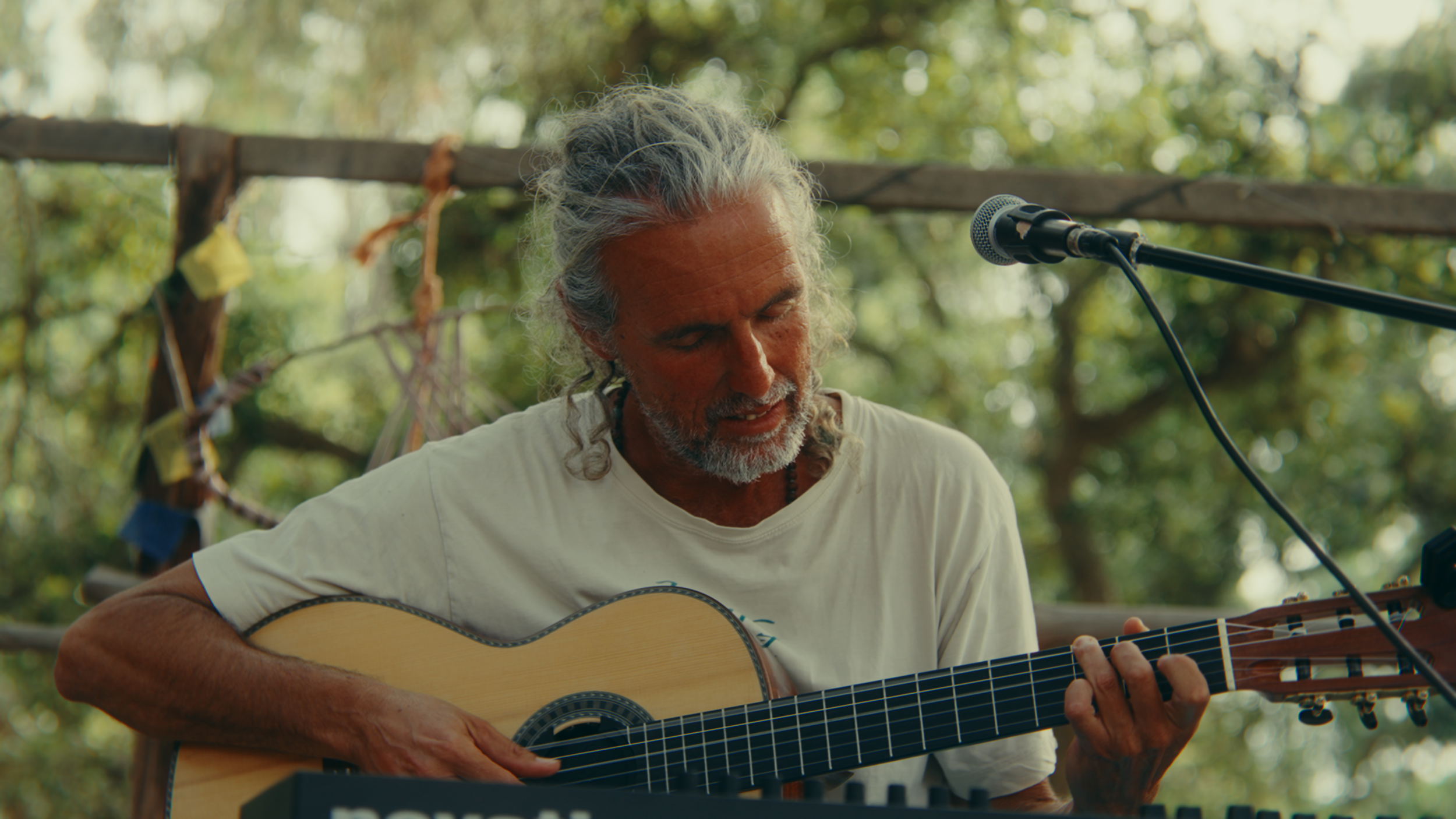 An older man with gray hair and a beard playing an acoustic guitar outdoors during daytime. There are green trees in the background, and a microphone is positioned in front of him.