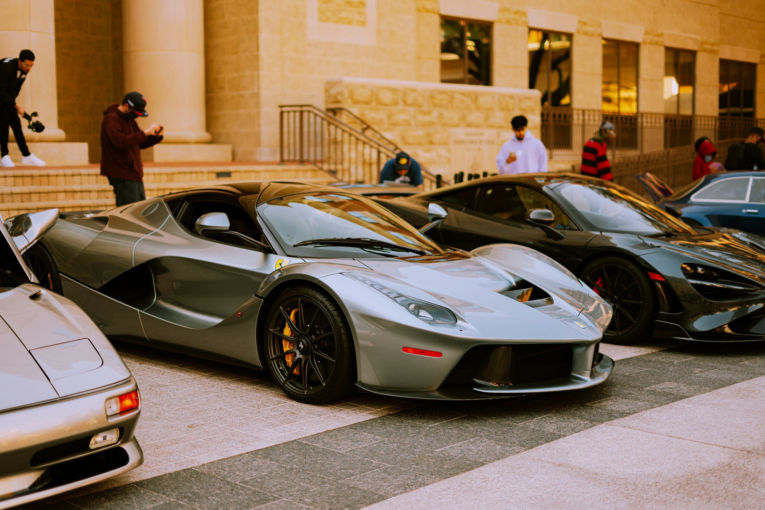 Multiple luxury sports cars, including a silver Ferrari, parked in front of a grand stone building with steps and large pillars, while several people are standing around, some taking photos or looking at their phones.