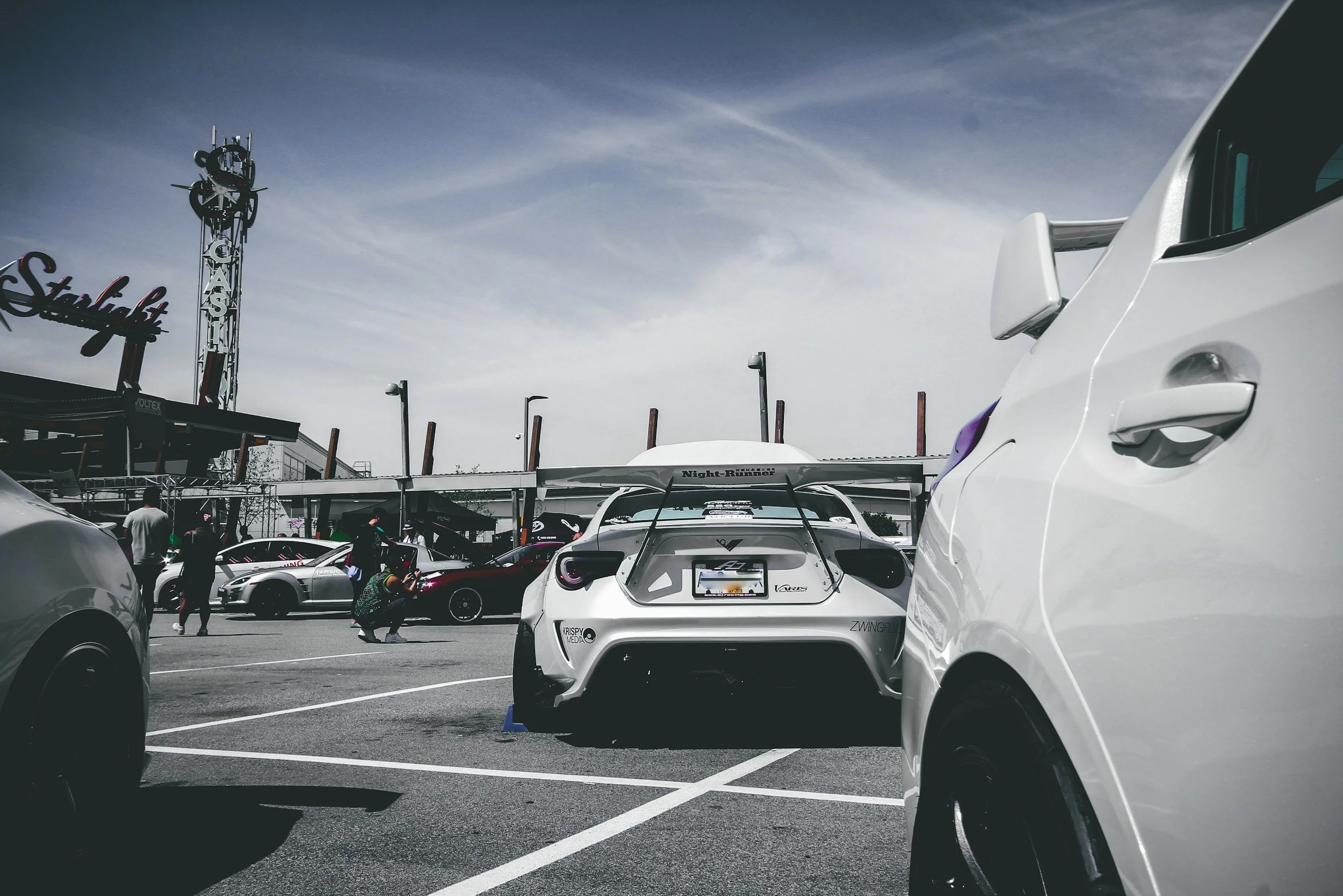 View of a parking lot filled with cars, including a white sports car with a large rear wing in the foreground, a person taking photos, and a large sign and tower with the word 'Starlight' and 'Circle K' in the background under a partly cloudy sky.