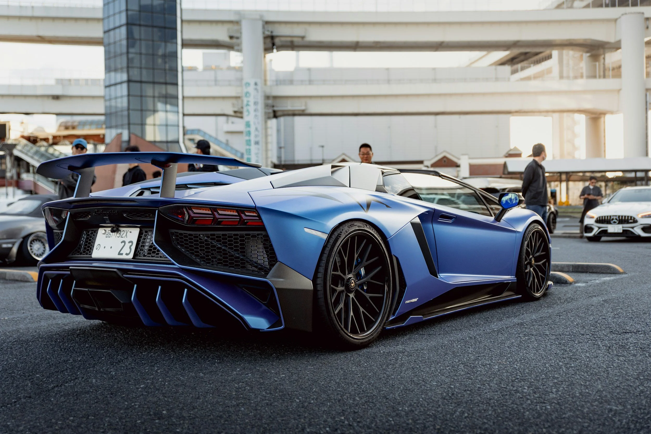 Blue Lamborghini sports car parked in an urban parking lot with several people and other vehicles in the background, under a multi-level highway overpass.