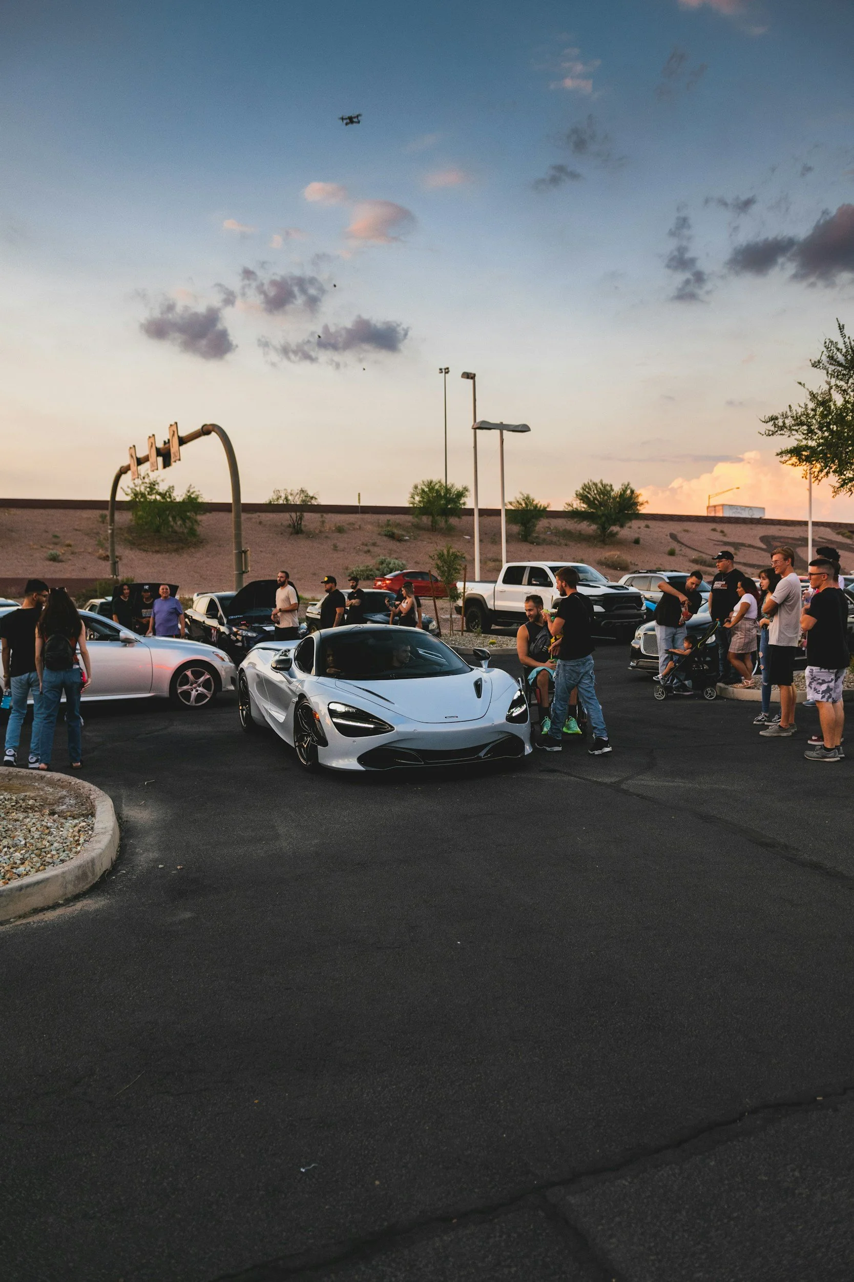 A group of people are gathered in a parking lot, engaging with three sports cars, including a silver convertible and a white sports car. The scene is during dusk, with a sky of scattered clouds and display lighting. There are trees and parked vehicles in the background.