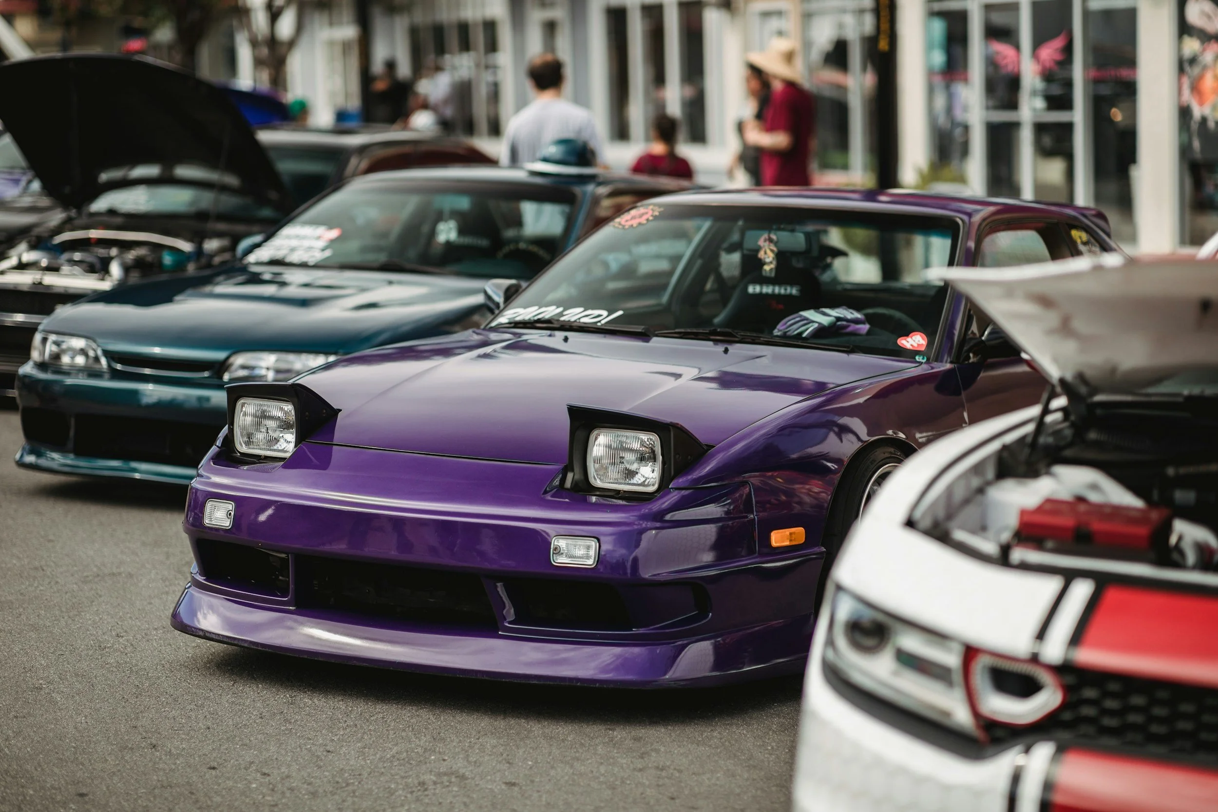 A row of compact sports cars, including a purple one in the foreground, parked on a street during daytime with some people walking in the background.