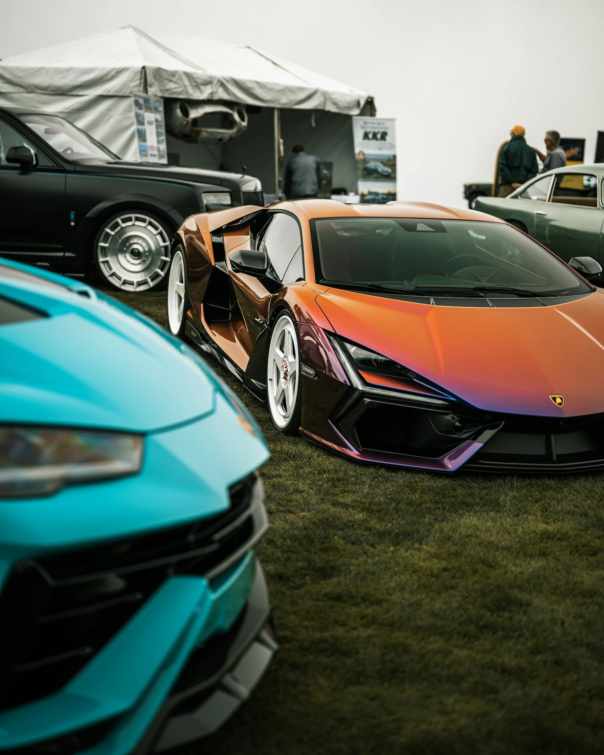 A lineup of luxury sports cars at an auto show, including an orange Lamborghini in the foreground, with other high-end cars and people in the background.