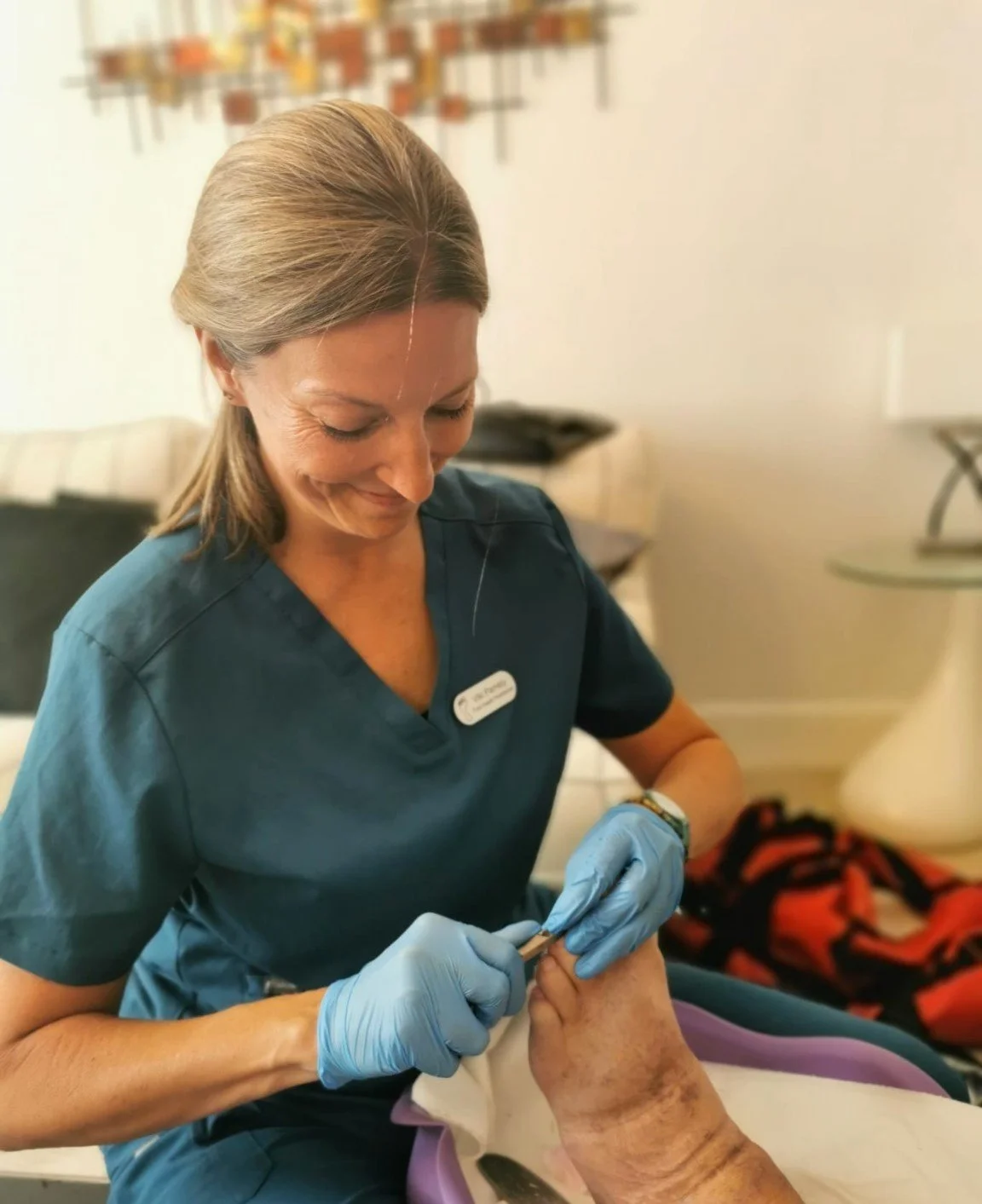 A healthcare worker in teal scrubs and blue gloves tending to a patient's hand, possibly giving injections, in a clinical setting.