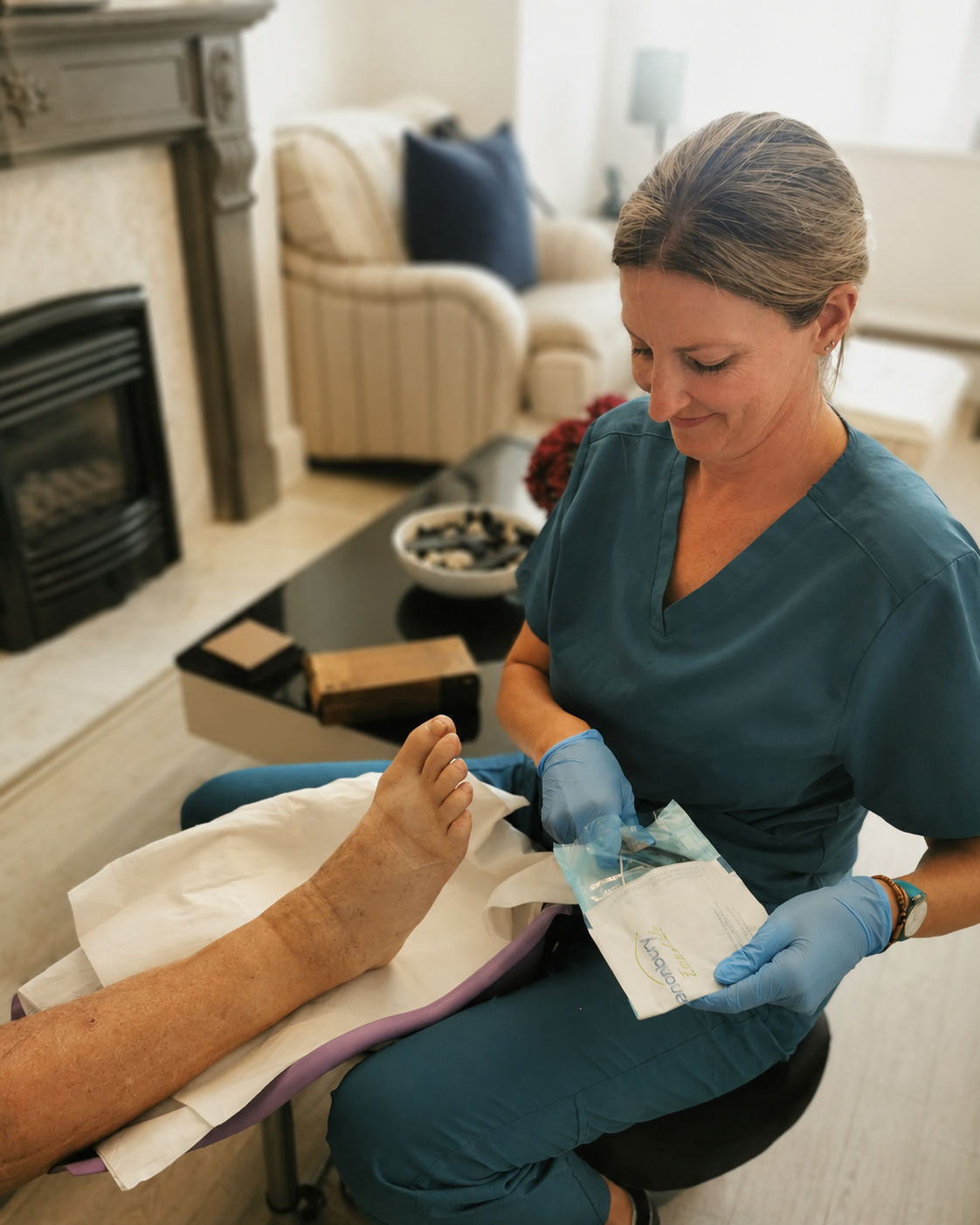 A healthcare worker, wearing blue scrubs and gloves, preparing to provide care or treatment to an elderly patient's foot, which is resting on a medical pad, in a cozy living room with a fireplace, sofa, and coffee table.