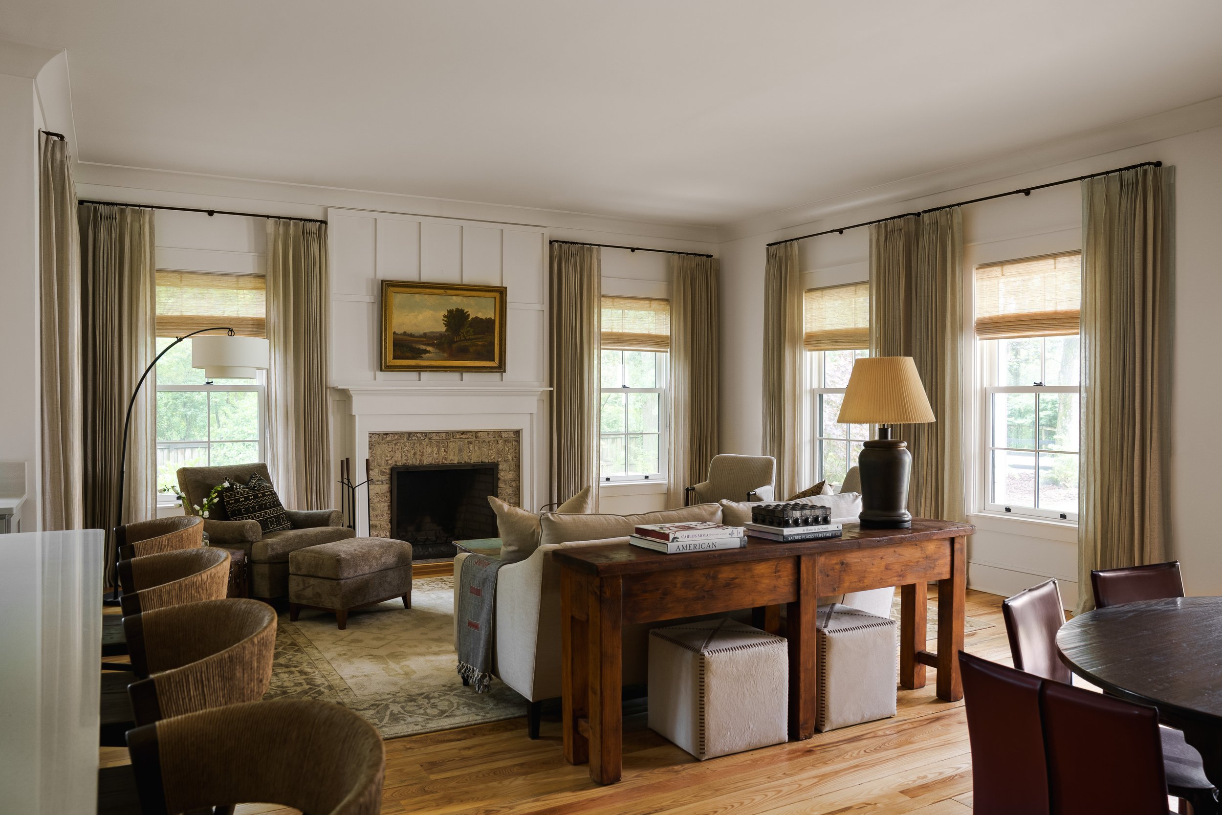 Living room with a fireplace, four large windows with blinds and curtains, a painting above the fireplace, a table with books and a lamp, and various chairs and couches.