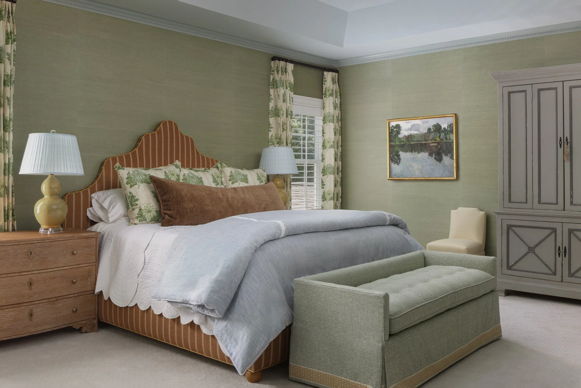 A bedroom with a green textured wall, a bed with a red and tan striped headboard, white bedding, brown and green pillows, and a light green upholstered bench at the foot of the bed. There are two nightstands with yellow lamps, a window with floral cu