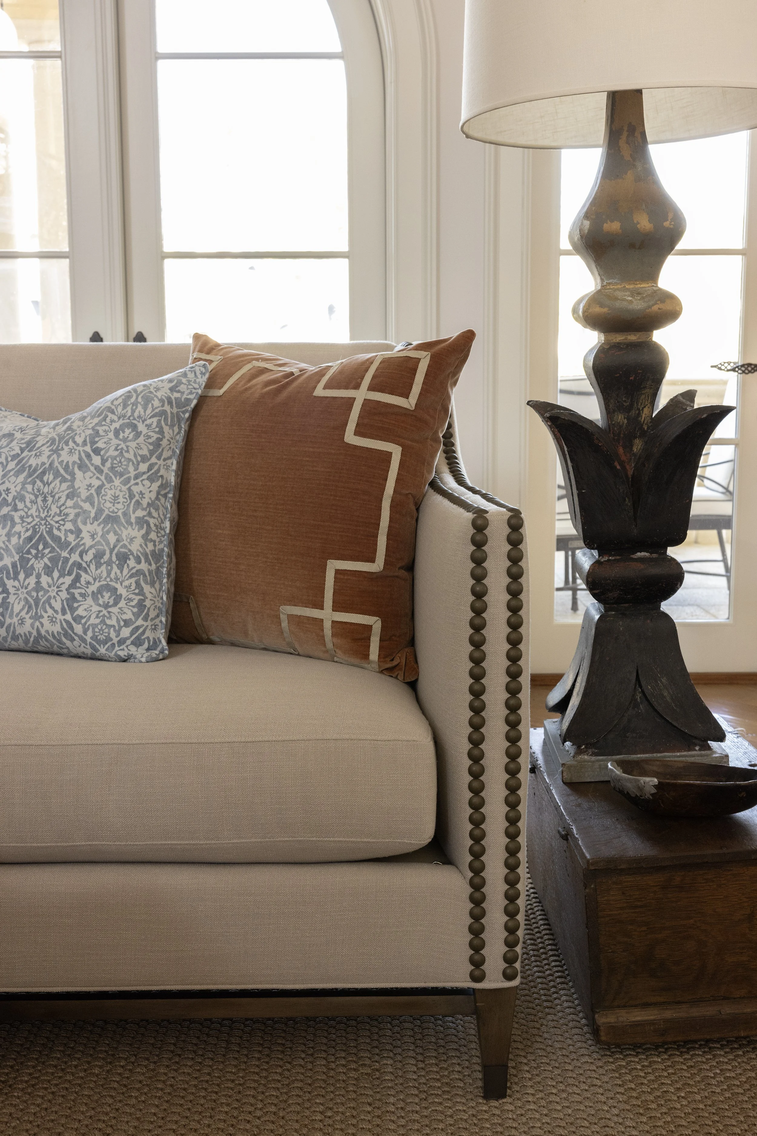 Close-up of a beige sofa with decorative pillows and an ornate wooden side table with a large table lamp, next to a window in a bright living room.
