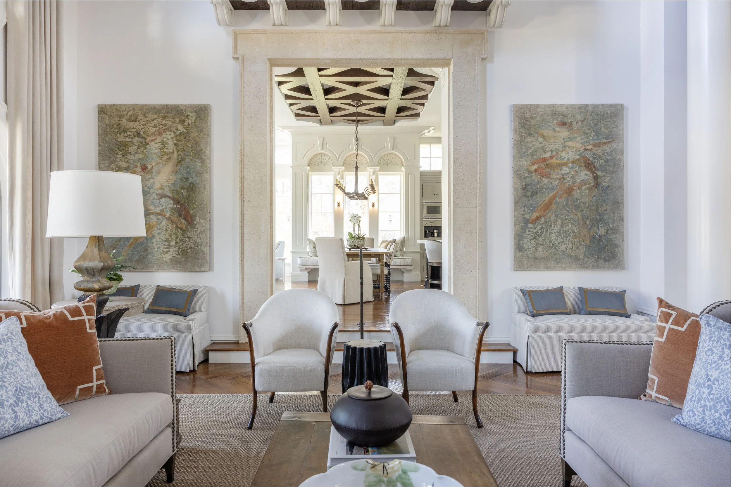 A view of a bright and spacious living room with cream-colored sofas, colorful pillows, a black stool, and a coffee table with a decorative plate. Two large paintings of fish hang on the wall, and a white lamp with a wooden base is placed next to the