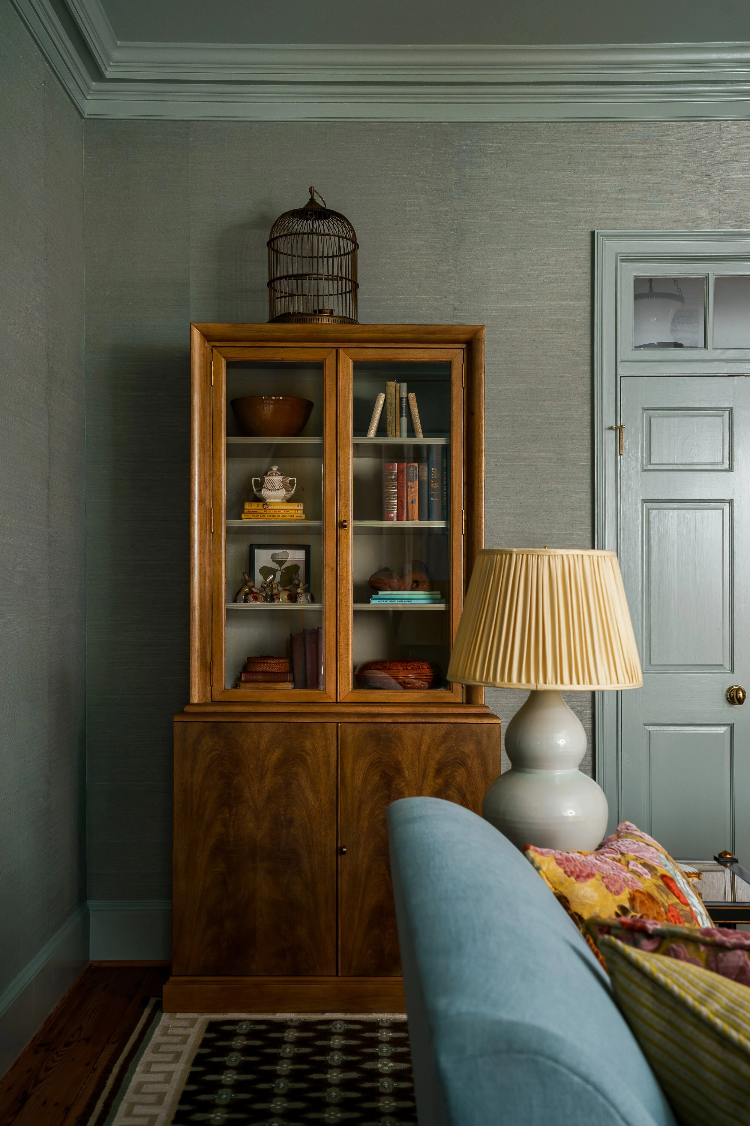 A living room corner with a wooden bookshelf, a white table lamp, and a blue sofa with a colorful pillow. The bookshelf has glass doors and contains books, decorations, and a teapot. The background features green walls, a door, and a birdcage on top of the bookshelf.