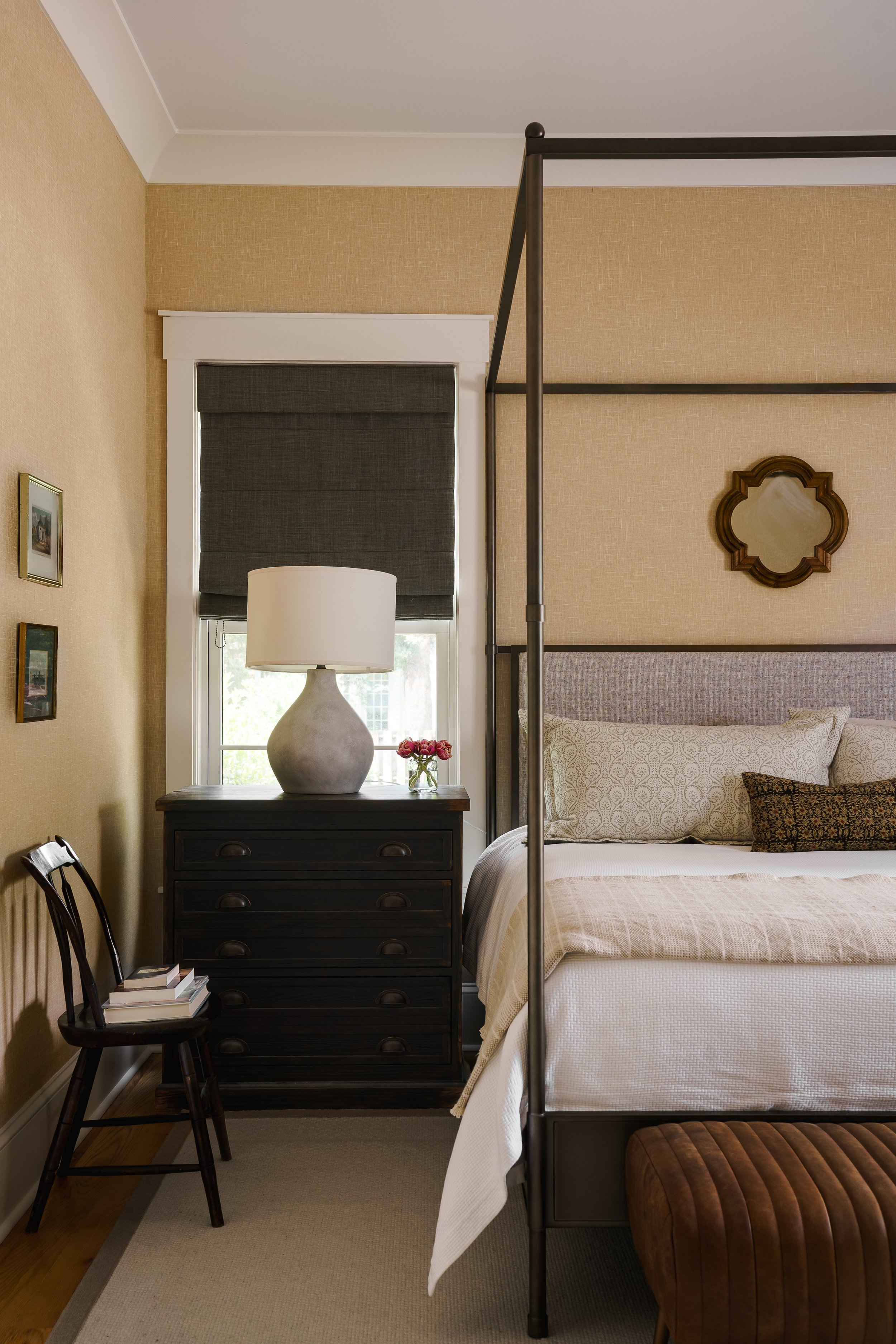 A bedroom with a bed, a black wooden dresser with a large white vase and a small pink flower arrangement, a window with gray Roman shades, part of a painting on the wall, and a small wooden chair with books on it. The bed has a beige comforter, a large patterned pillow, and a dark brown leather ottoman