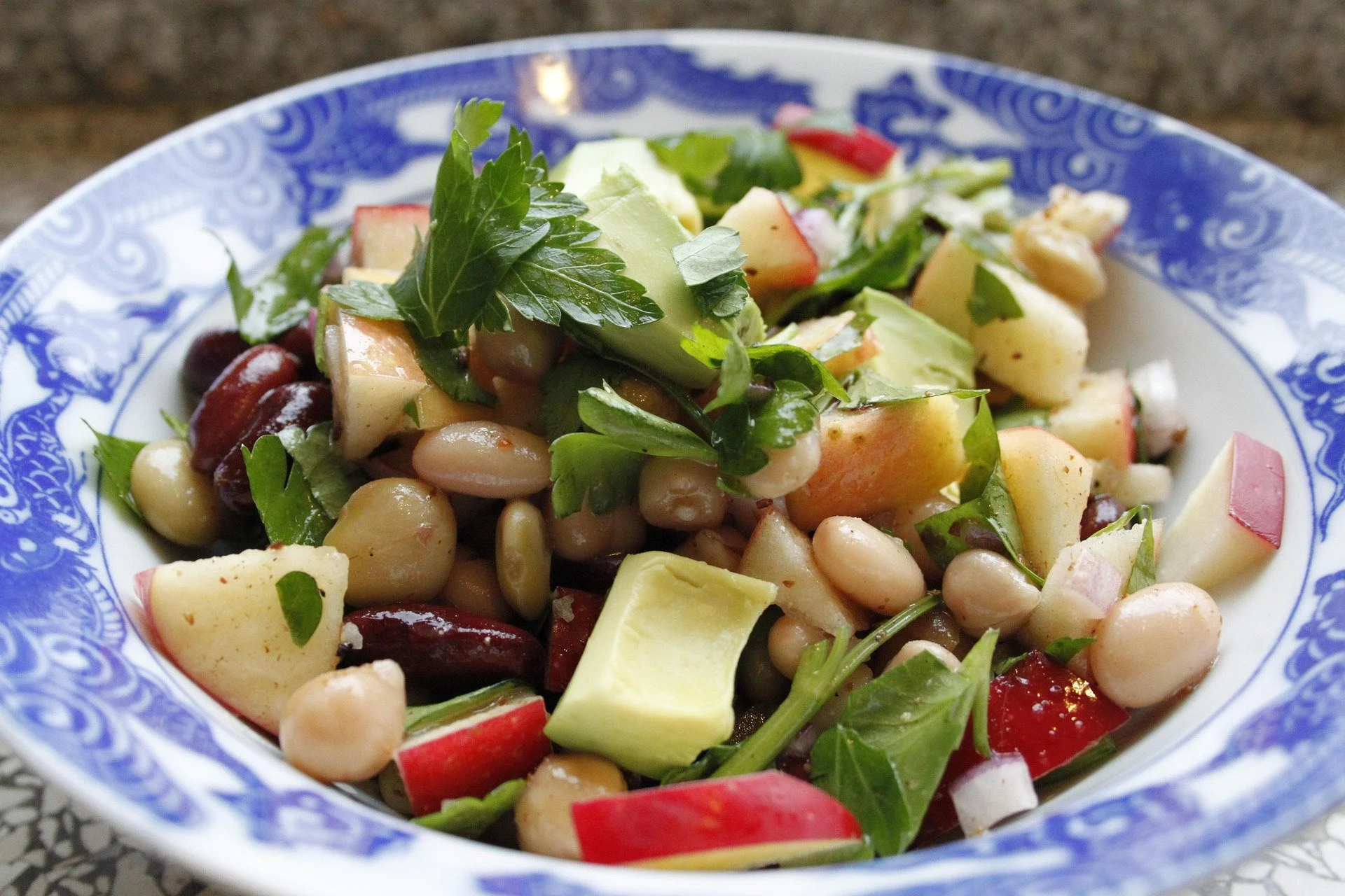 A fresh bean salad with diced apples, cubed avocado, and parsley served in a blue and white decorative bowl.
