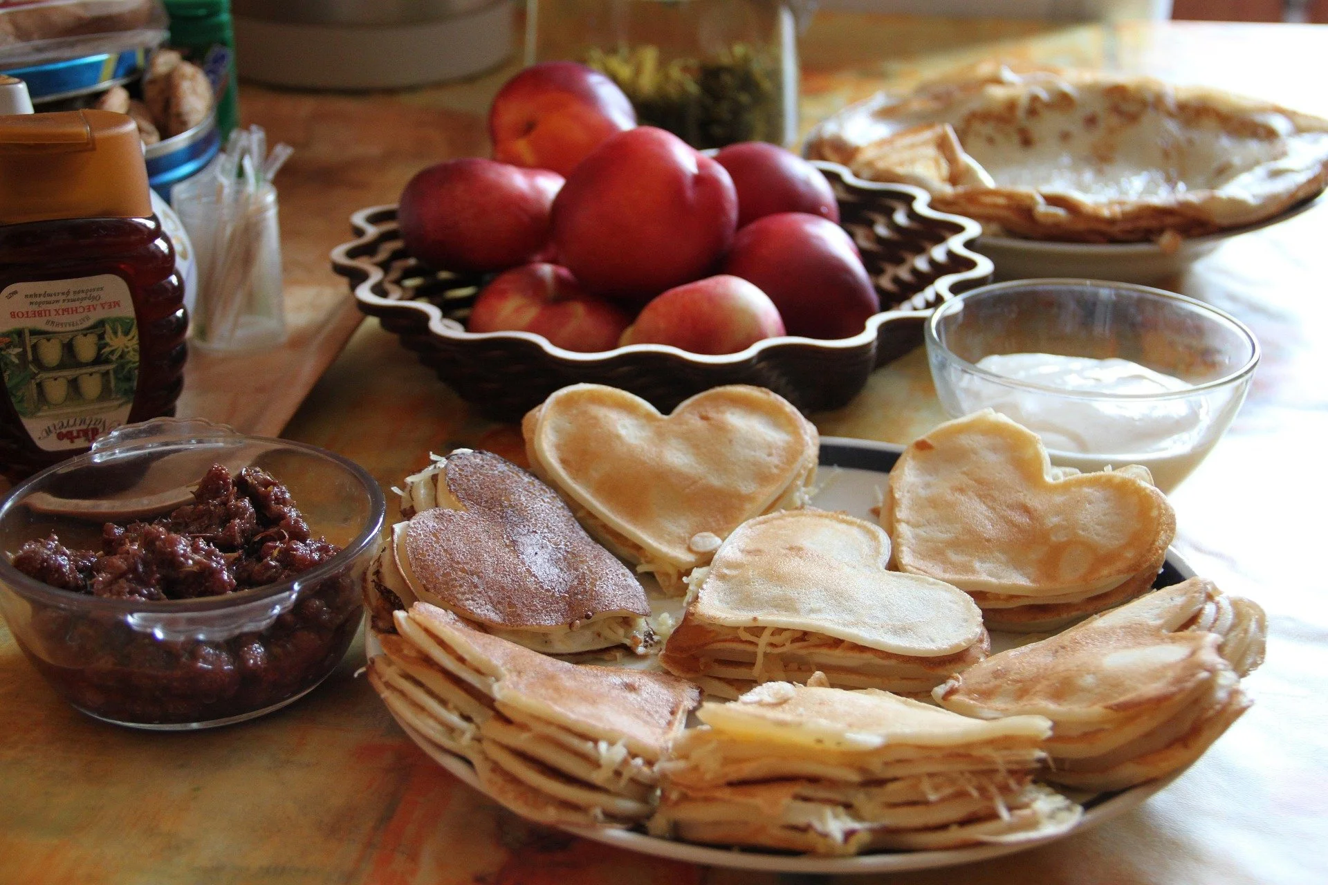 A breakfast spread featuring heart-shaped pancakes, fresh nectarines or plums in a bowl, honey, jam, and yogurt.