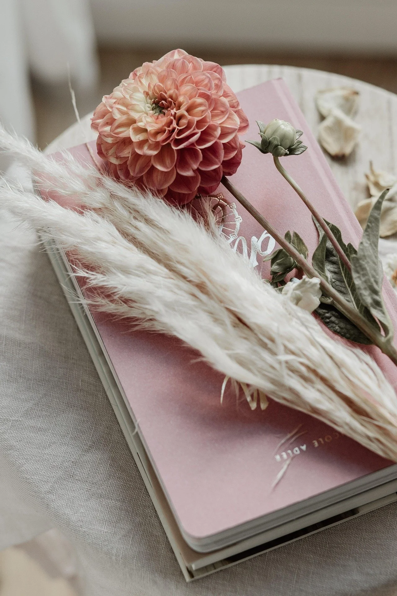 Close-up of a pink dahlias flower, a budding flower, book with a pink cover, fluffy white pampas grass, and dried flower petals on a pink notebook.