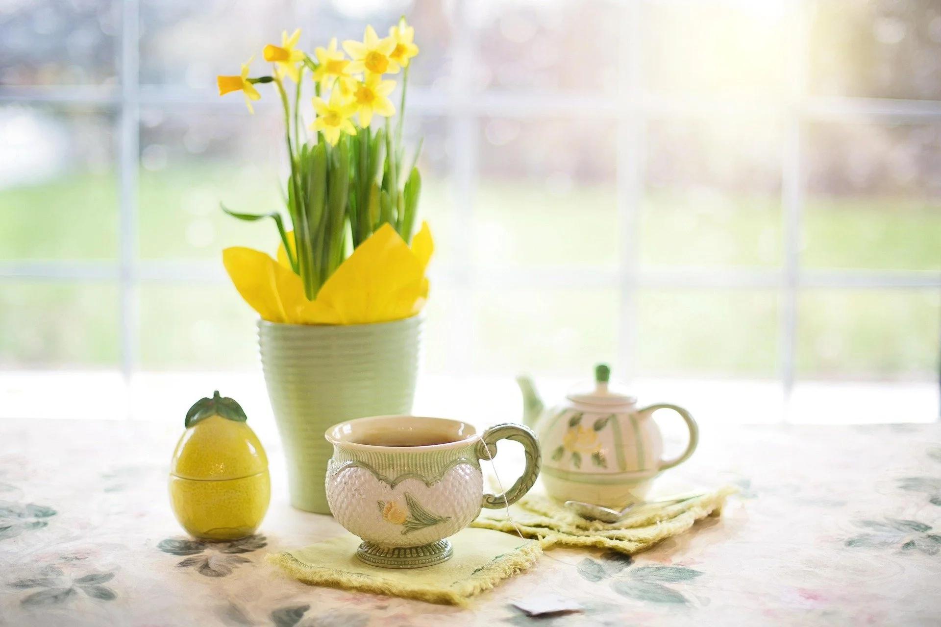 Vase of yellow daffodils on a table with teacups, a lemon-shaped container, and a teapot in the background, with a bright window behind.