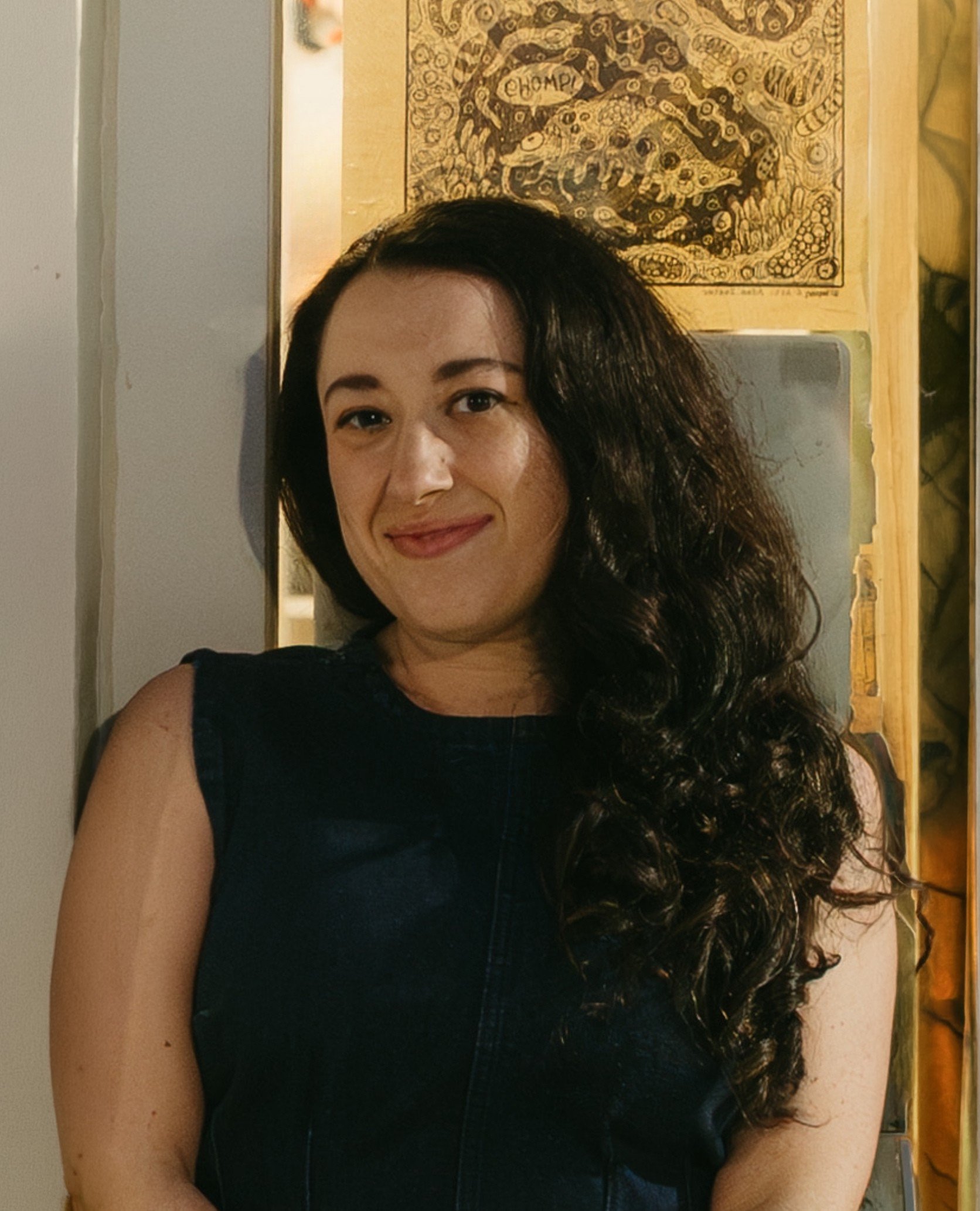 Marissa Finkelstein, the Program Manager of Arts & Well-Being at the New Jersey Performing Arts Center, wearing a black sleeveless top, standing in front of a wall decorated with a black and gold painting.