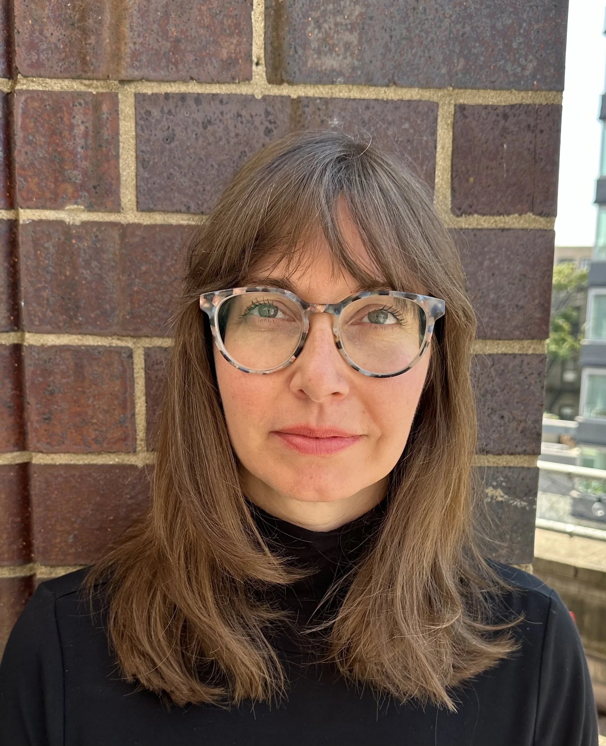 Lis Chirinos, the Director of Arts in Public Health at Sing for Hope, wearing glasses standing in front of a brick wall.