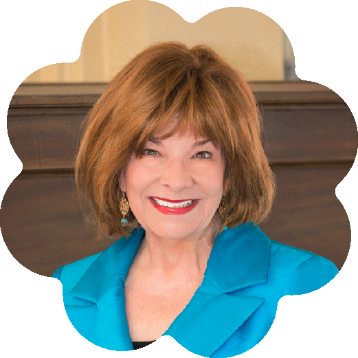 Margery Pabst Steinmetz, Author, Philanthropist and Co-Founder of the Pabst Steinmetz Foundation, wearing a bright blue blazer and earrings, smiling in an indoor setting with wooden paneling in the background.