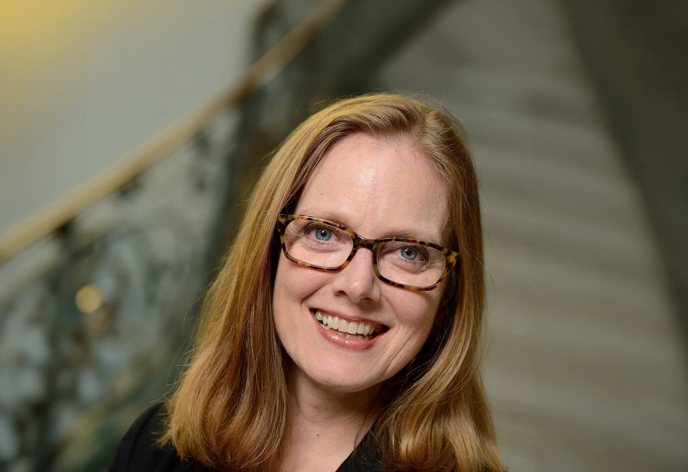 Sarah Hoover, the Associate Dean for Innovation in the Arts and Health at The Peabody Institute of the Johns Hopkins University, wearing glasses, smiling, indoors with blurred background of a staircase.
