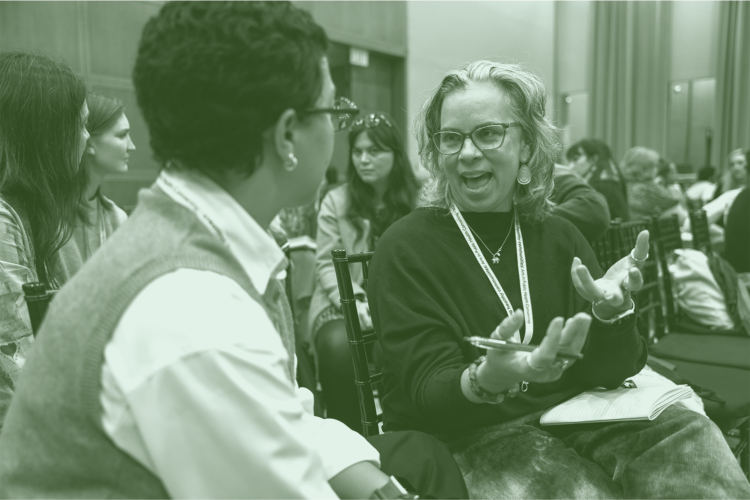A woman with glasses and curly hair engaged in a conversation, gesturing with her hands, during a conference or meeting. She has a notebook on her lap and is wearing a lanyard. Several other people are seated in the background, listening and participating.
